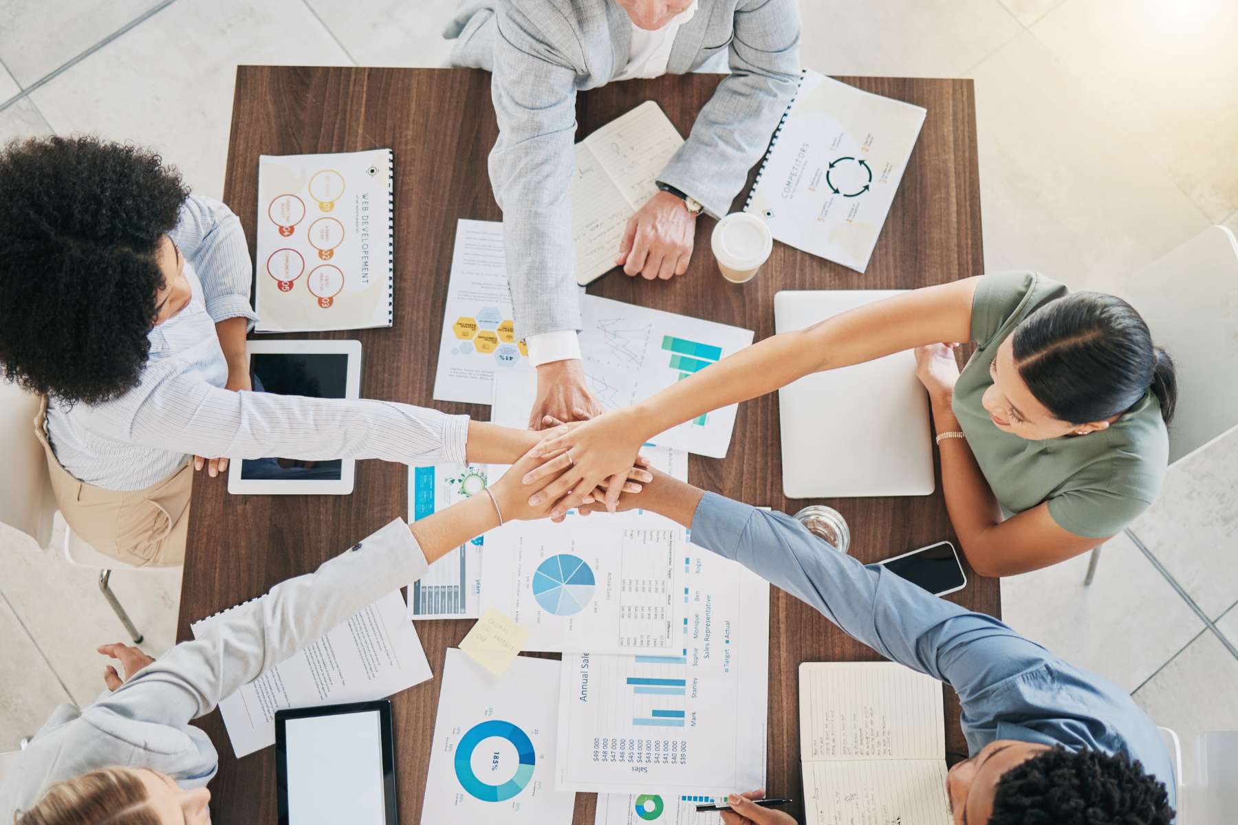 Eine Gruppe junger diverser Büroangestellter wird aus der Vogelperspektive gezeigt, während sie an einem großen Schreibtisch sitzt auf dem viele Papiere liegen, und die Hände als Teamgeste in der Mitte aufeinanderlegen.