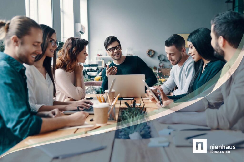 Eine gruppe junger Büroangestellter sitzt als Team um einen großen Holztisch herum und unterhält sich angeregt, während Laptops und Schreibutensilien auf dem Tisch liegen.