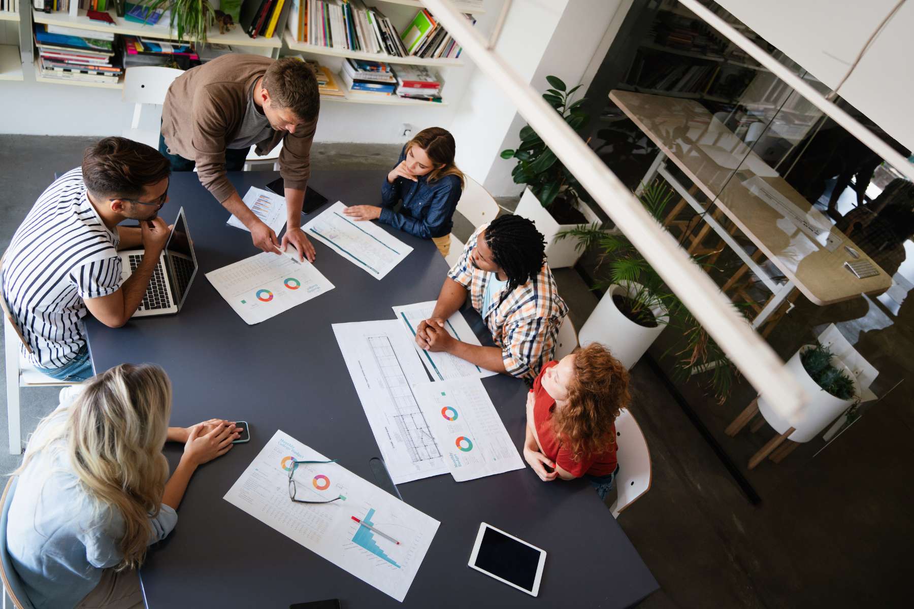Eine Gruppe diverser Büroangestellter ist aus der Vogelperspektive dabei zu sehen, wie sie am Tisch sitzt und ein Projekt bespricht.