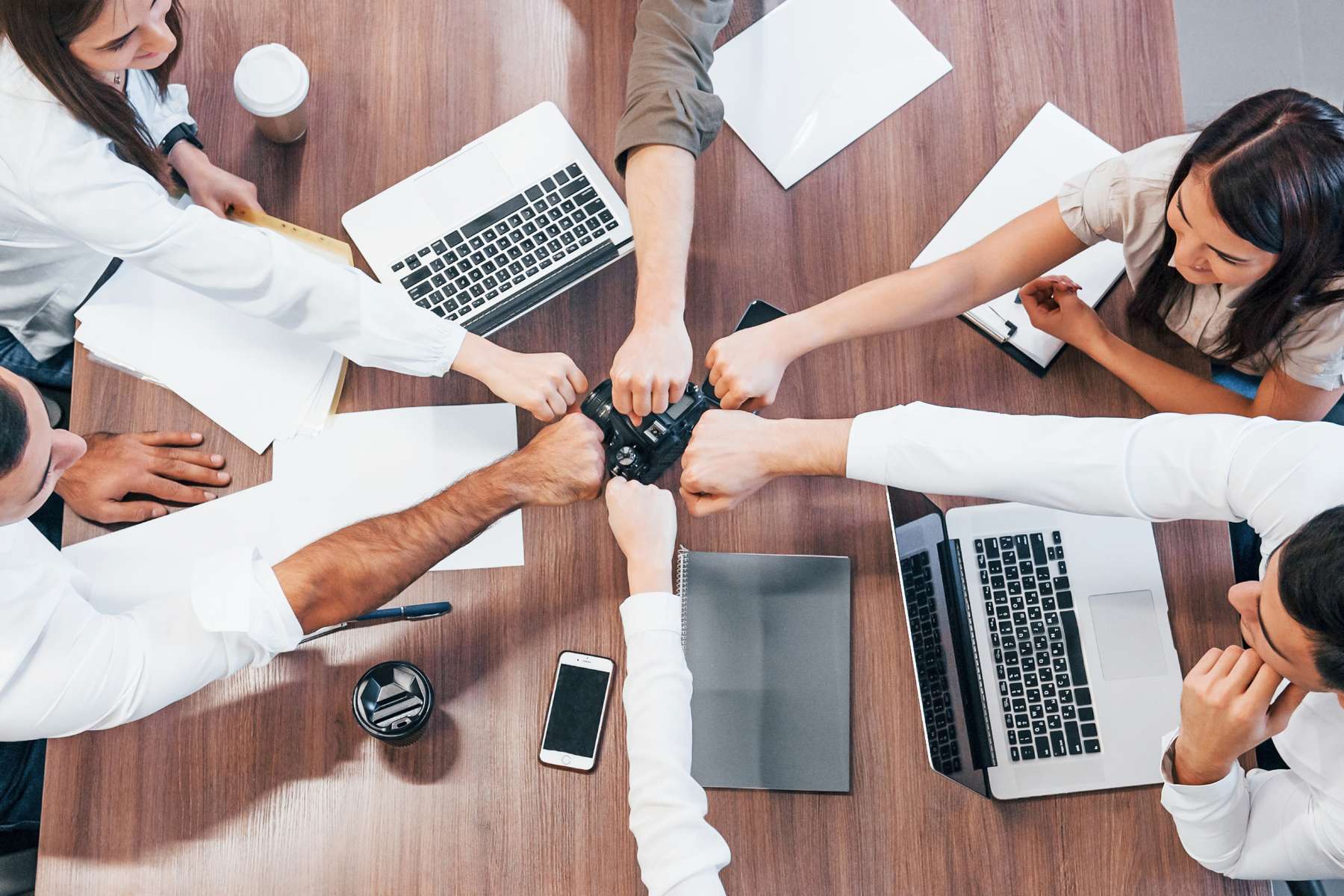 Eine Gruppe Büroangestellter sitzt um einen großen Holztisch herum und streckt sich über den Tisch hinweg lächelnd die Fäuste entgegen, als Sinnbild für Teamwork und Arbeiten ohne Chefstruktur.