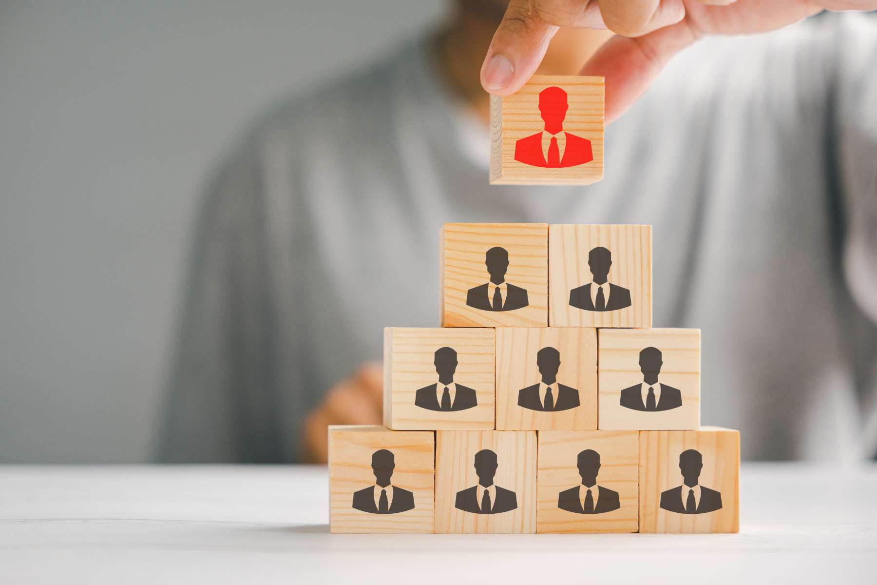 Mehrere Bauklötze aus Holz stehen zu einer Pyramide gestapelt auf einem Tisch, während die unteren Reihen Silhouetten eines Mannes im Anzug in schwarz zeigen, und die oberste Silhouette rot ist., als Sinnbild für klassische Chefstrukturen.