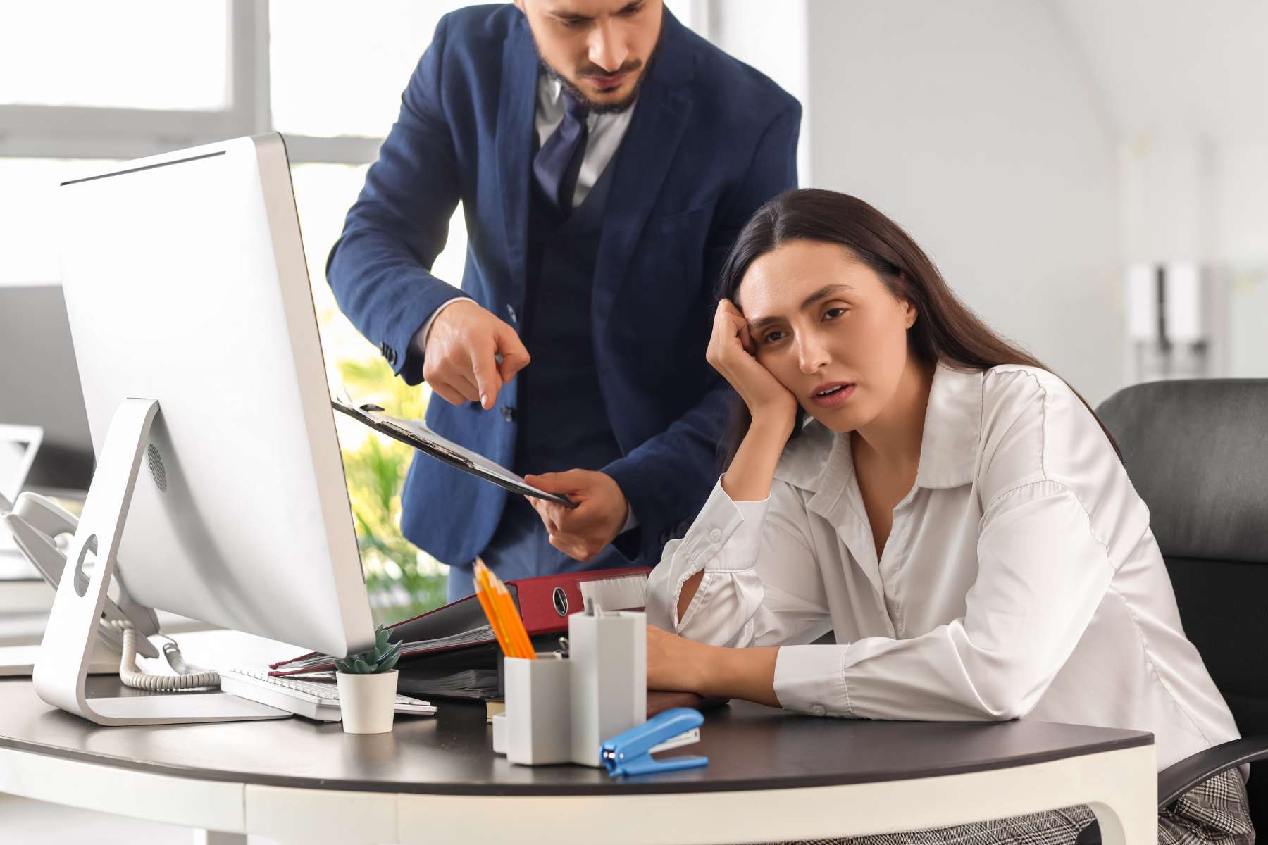 Eine genervte Angestellte sitzt an einem Schreibtisch vor einem Bildschirm und dreht sich genervt von einem Kollegen weg, der neben ihr steht und ihr etwas auf einem Klemmbrett zeigen will.