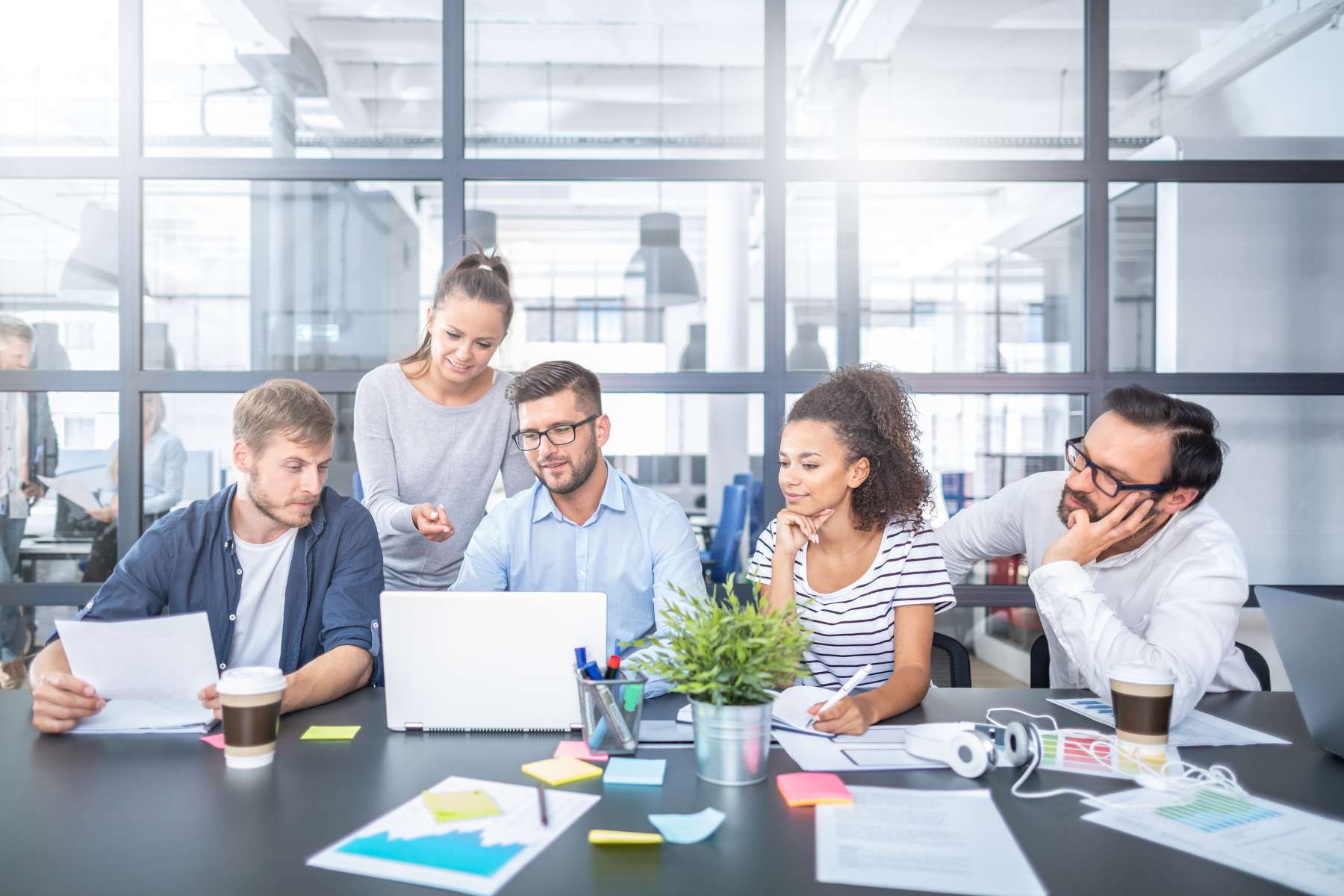 5 junge diverse Angestellte sitzen bzw. stehen im Büro an einem großen Tisch und schauen sich etwas auf einem Laptop an.