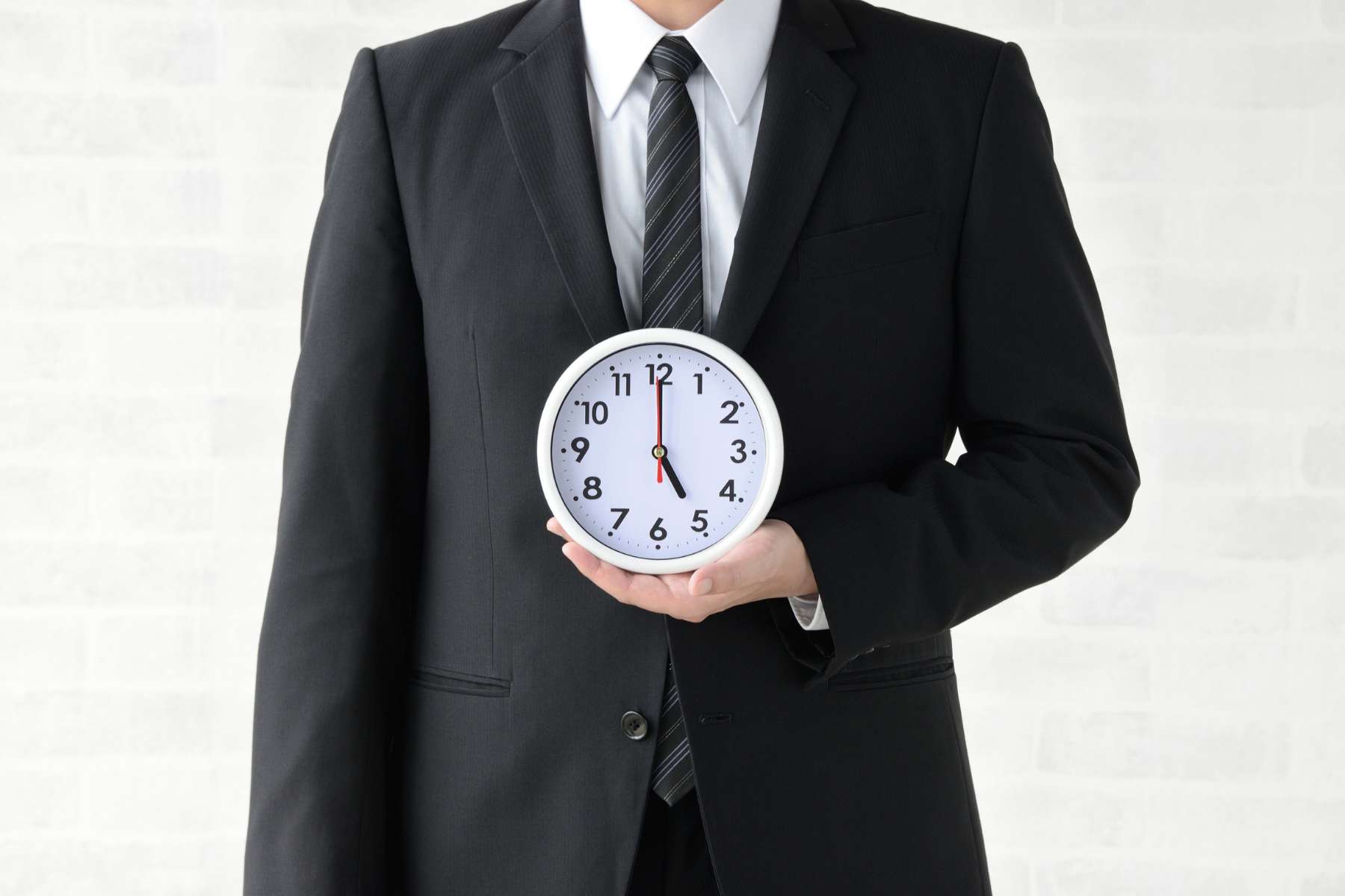 Ein Mann in schwarzem Anzug, von dem man nur den Oberkörper ohne Kopf sieht, hält eine weiße Uhr in der linken Hand vor seine Brust.