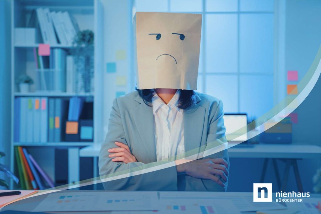 Büromitarbeiterin mit verschränkten Armen am Schreibtisch und Papiertüte mit unglücklichem Gesicht auf dem Kopf.