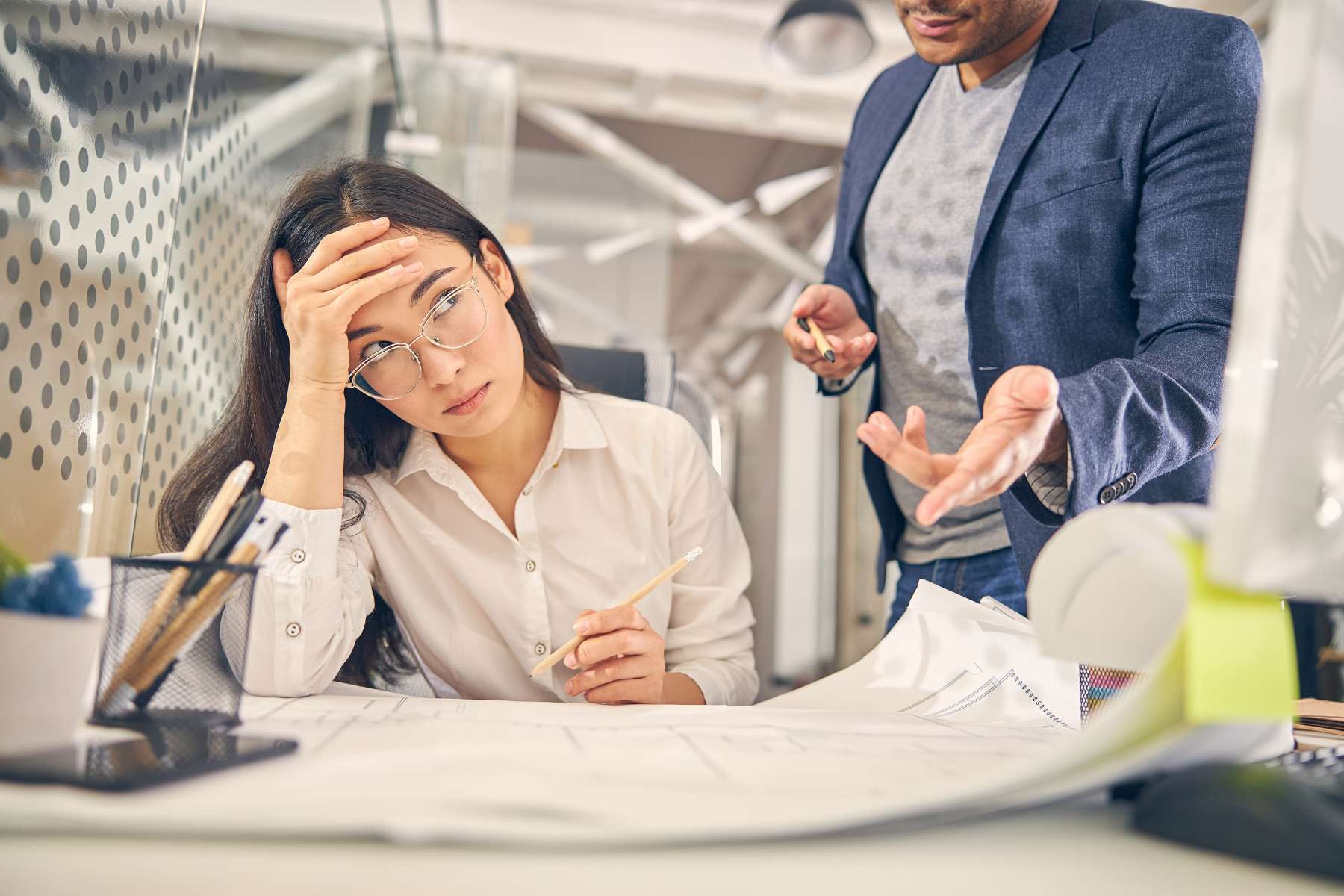 Eine Büroangestellte sitzt am Schreibtisch und schaut genervt zur Seite, von wo ein Kollege auf sie einredet.
