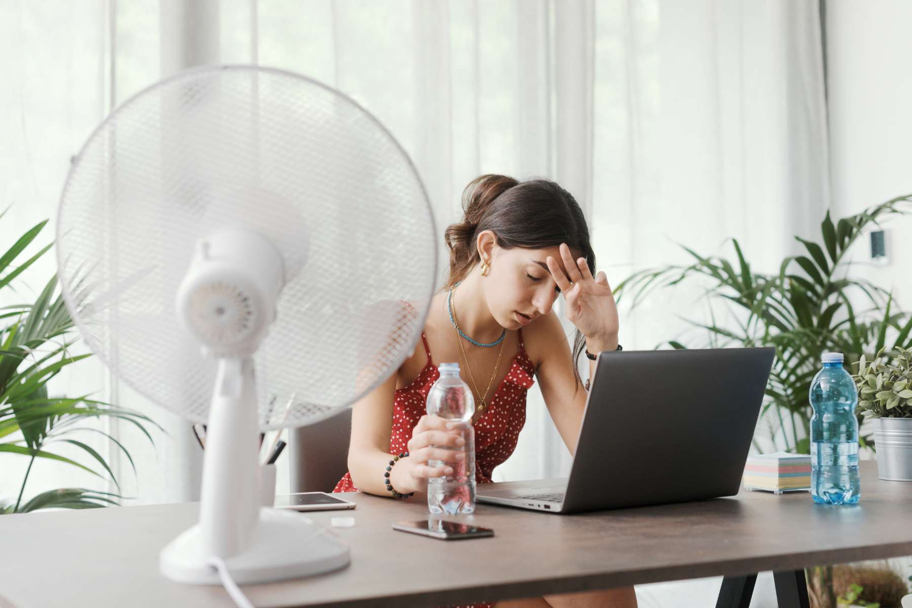 Eine Frau sitzt mit einer Flasche Wasser in der Hand schwitzend am Schreibtisch, während ein Ventilator vor ihr steht.