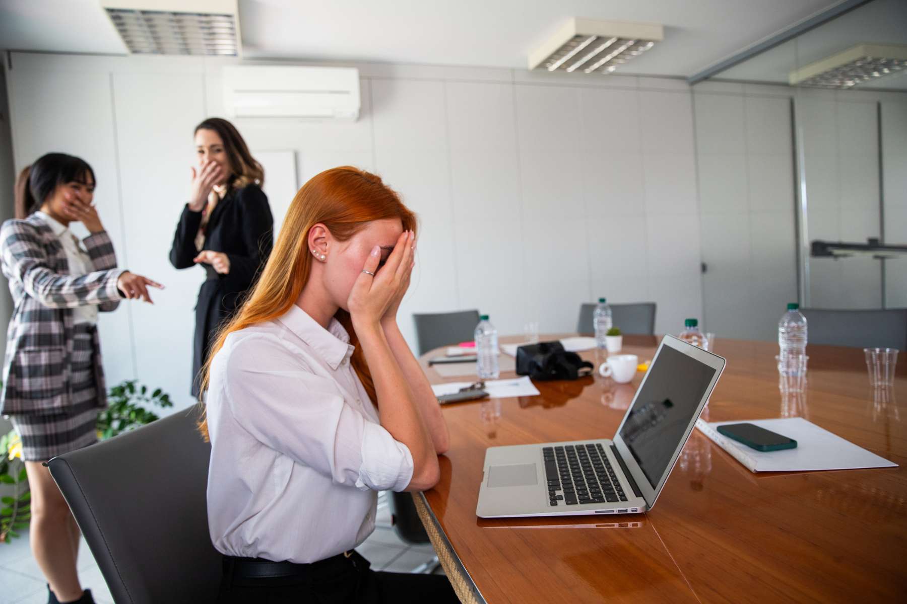 2 Büromitarbeiterinnen machen sich über eine am Schreibtisch sitzende Kollegin lustig, die die Hände vor dem Gesicht zusammenschlägt