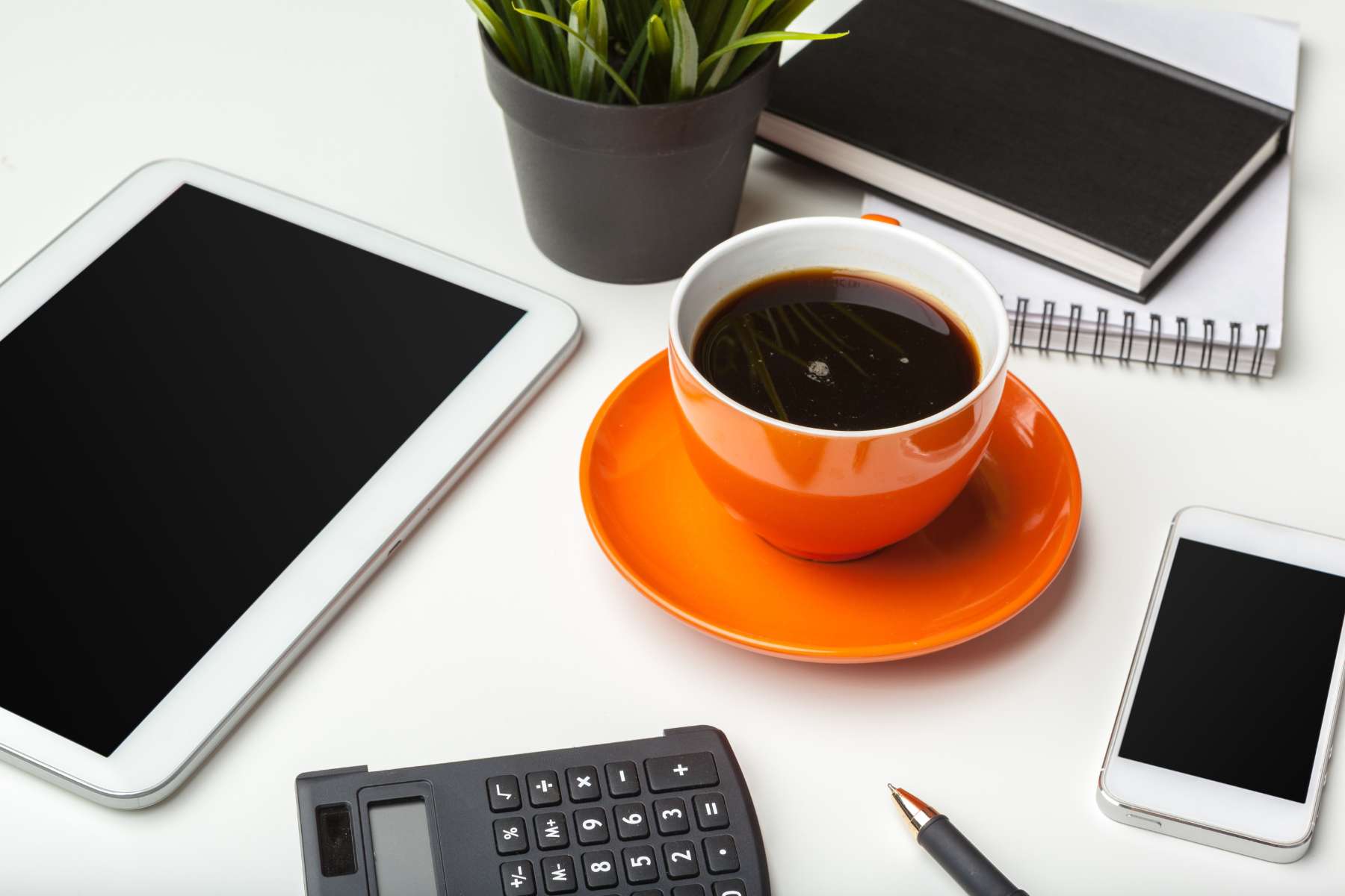 Eine orangene Kaffeetasse steht auf einem Schreibtisch, drumherum liegt ein Tablet, ein Handy, ein Taschenrechner und weitere Büroutensilien.