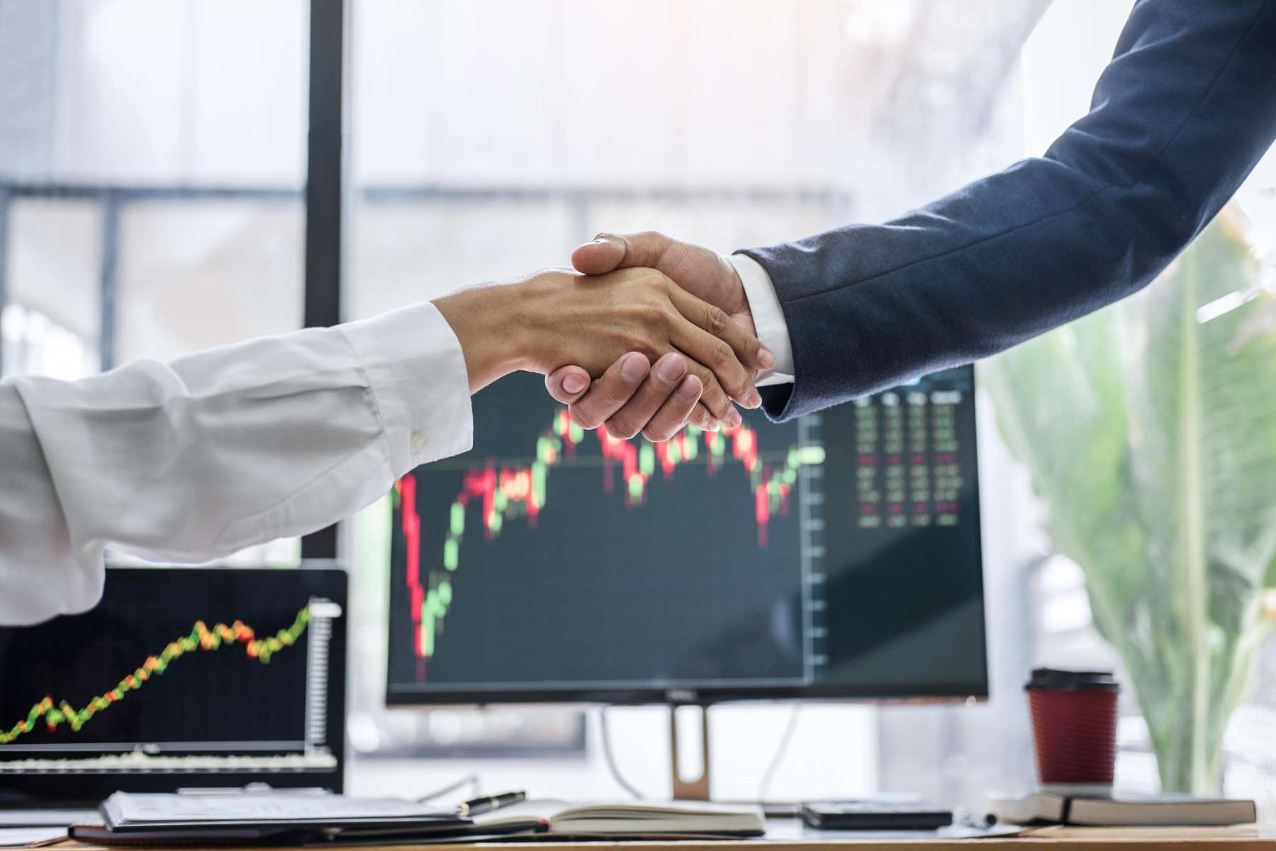 Eine weibliche Angestellte und ein männlicher Angestellter schütteln sich die Hand, während im Hintergrund auf zwei Bildschirmen unscharfe Statistiken zu sehen sind.