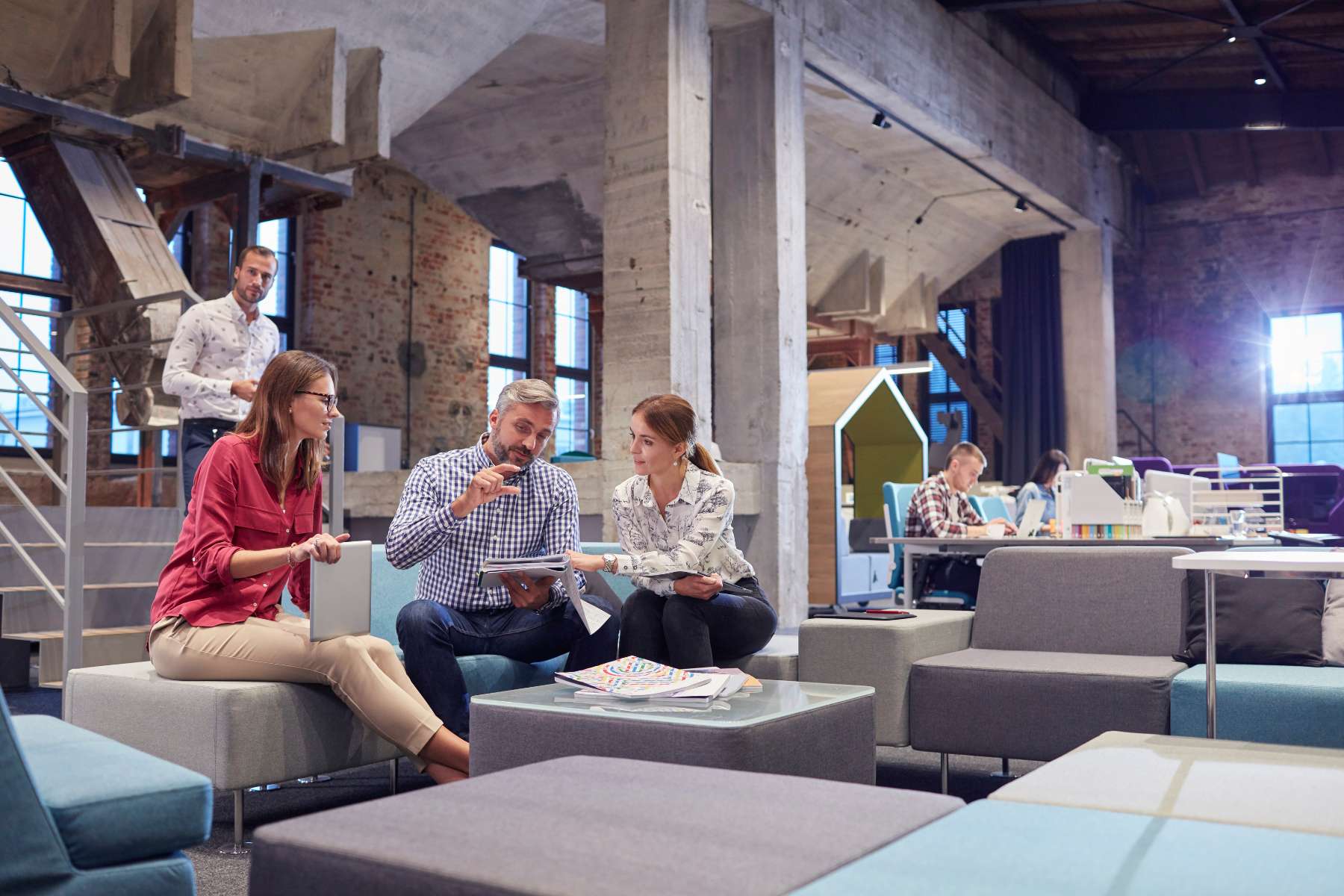 Drei Büroangestellte sitzen in einem Loungebereich eines Büros, während im Hintergrund ein weitläufiger Bereich zu sehen ist, in dem andere Angestellte an Schreibtischen arbeiten.