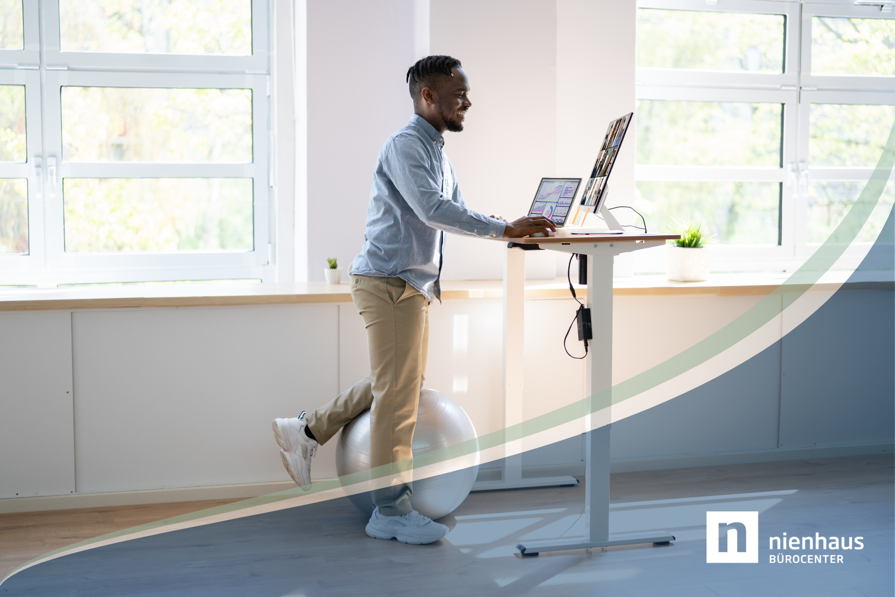 Junger Mann arbeitet im Büro an einem höhenverstellbaren Schreibtisch und stütz dabei ein Bein auf einen Gymnastikball.
