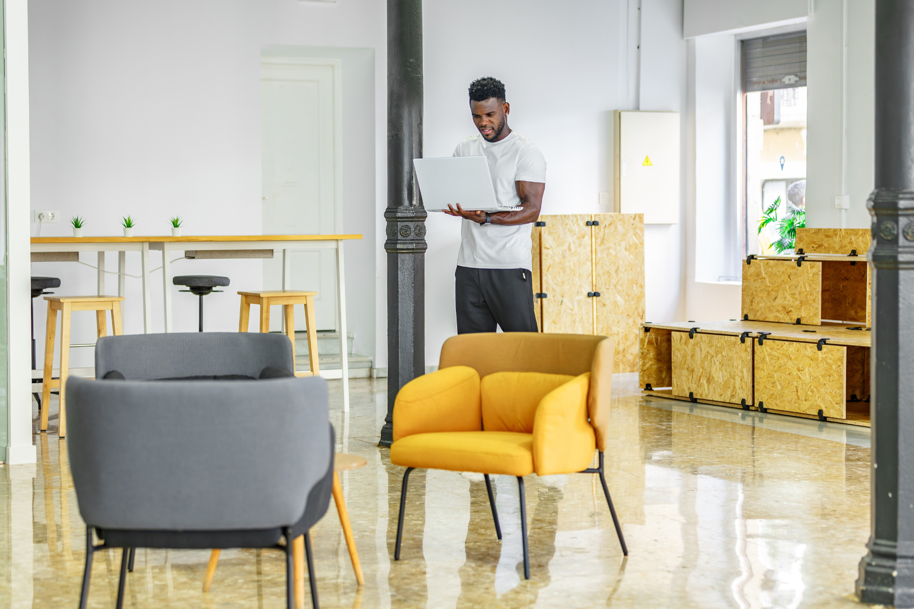 Junger Angestellter mit Laptop in der Hand steht in stylischem Büroraum mit grauen und gelben Stühlen, Boxen aus Holz auf der rechten Seite und Stehtischen plus Stehhockern im Hintergrund.