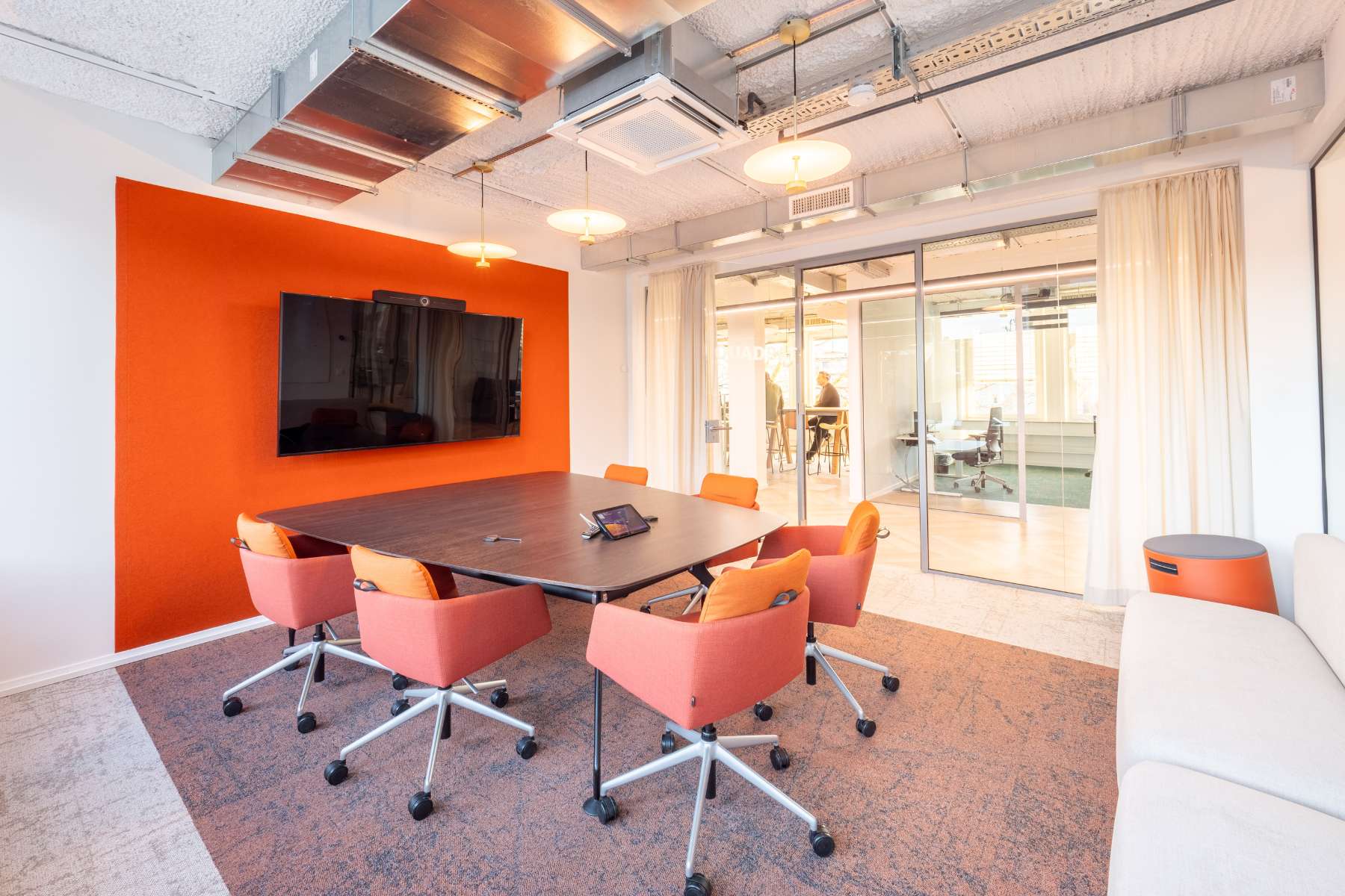 Abtrennbarer Besprechungsraum inklusive Holztisch, orangenen Stühlen, farblich abgestimmter Wand, weißer Couch und großem, an der Wand befestigtem Monitor.