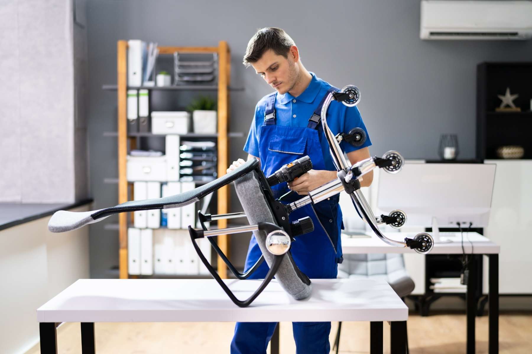 Ein Handwerker in blauem Overall repariert einen grauen Bürostuhl, den er auf einem weißen Schreibtisch abgelegt hat.
