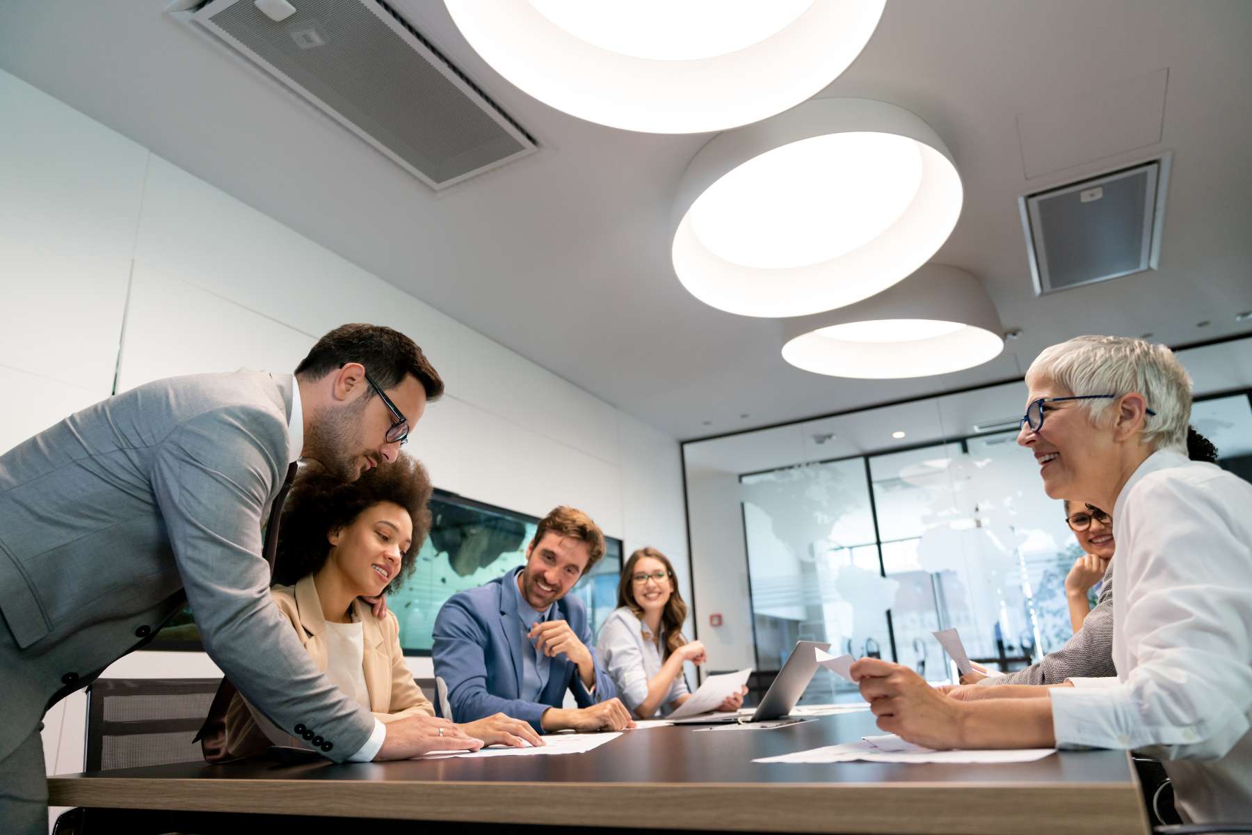 Eine Gruppe von sich unterhaltenden Büroangestellten sitzt an einem langen Tisch über dem drei große Deckenlampen im Stil von Smart Lighting angebracht sind.