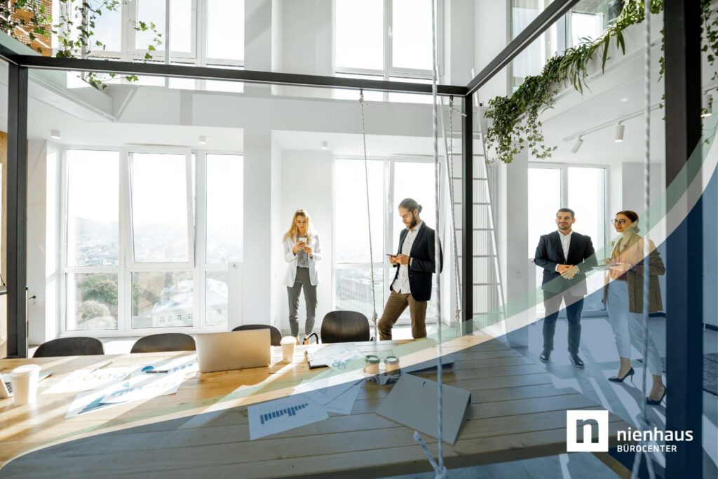 Vier Mitarbeiter stehen in einem hellen Büro mit großer Glasfront, hohen Decken und modernem Holztisch im Vordergrund,