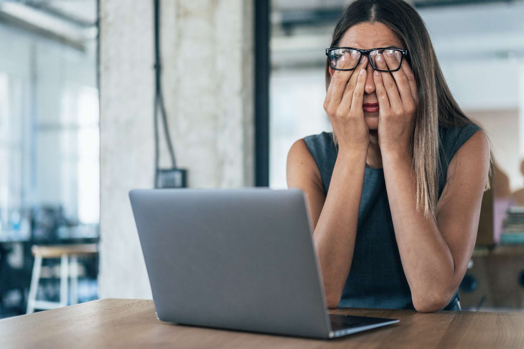 Eine Frau sitzt vor ihrem Laptop an einem Schreibtisch und hält sich erschöpft die Hände vor ihr Gesicht.