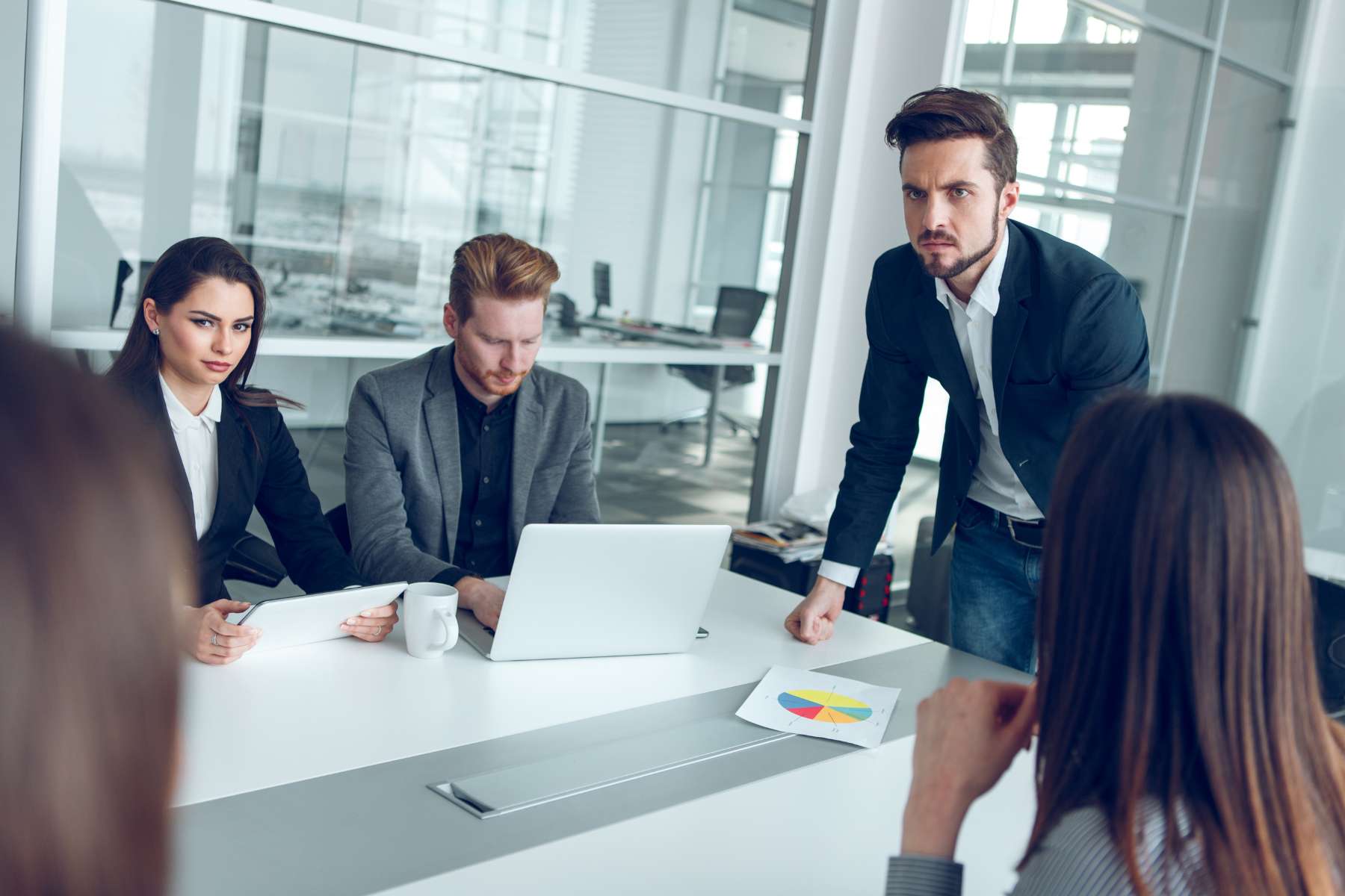 Vier Büroangestellte sitzen an einem weißen Tisch, während sich ihr stehender Vorgesetzter verärgert auf den Tisch lehnt.