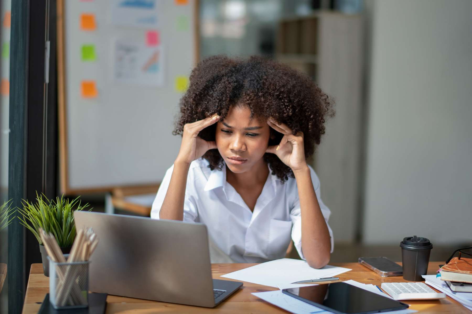 Eine junge schwarze Frau sitzt an einem Schreibtisch vor einem Laptop und vielen Papieren und hält sich mit gestresstem Gesicht die Hände an die Schläfen.