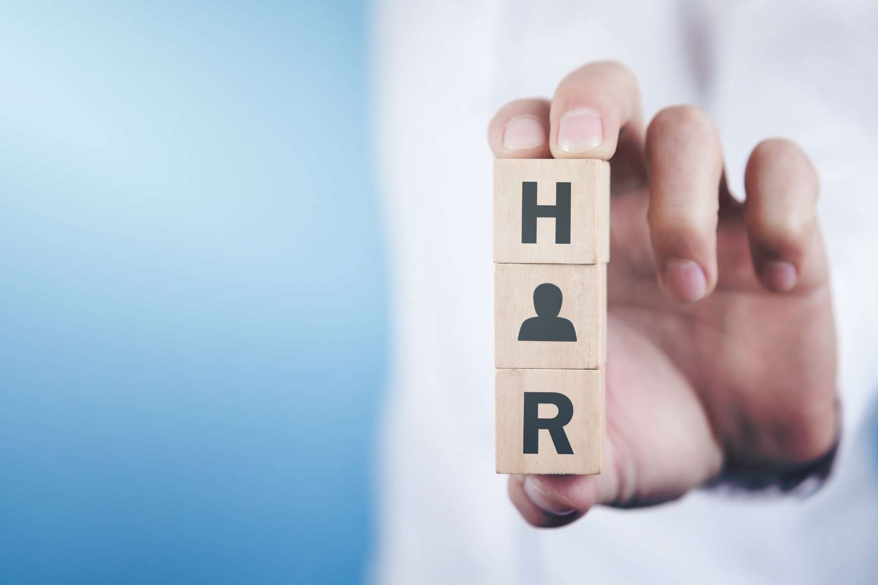 Ein Mann hält drei Holzbausteine zwischen zwei Fingern. Auf dem oberen steht der Buchstabe H, in der Mitte ist eine Silhouette einer Person abgebildet und auf dem unteren steht der Buchstabe R, als Zeichen für das Thema Human Ressources.