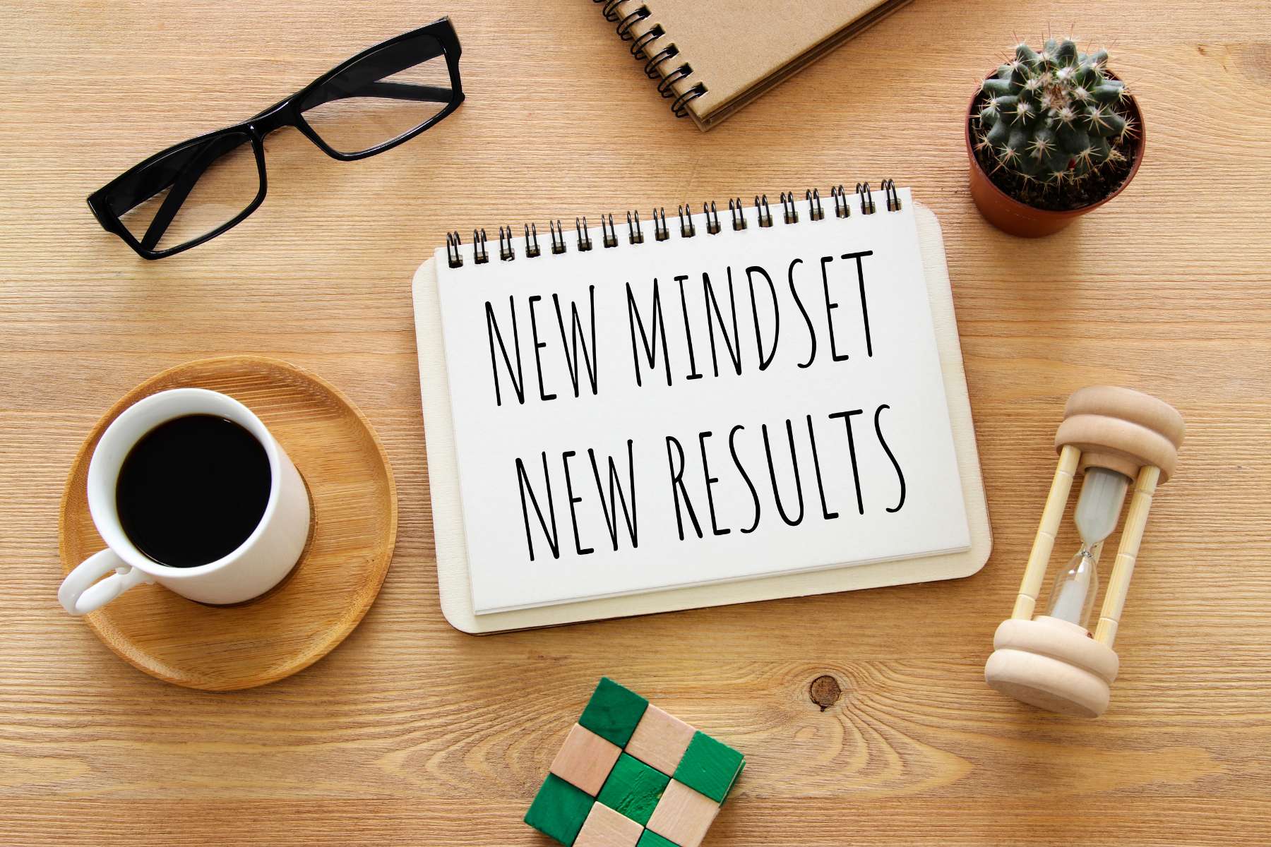 Ein Holztisch von oben, auf dem eine Tasse Kaffee steht, eine Sanduhr, eine Brille und ein Zauberwürfel liegt und mittig ein Block mit den Worten New Mindset New Results zu sehen ist.
