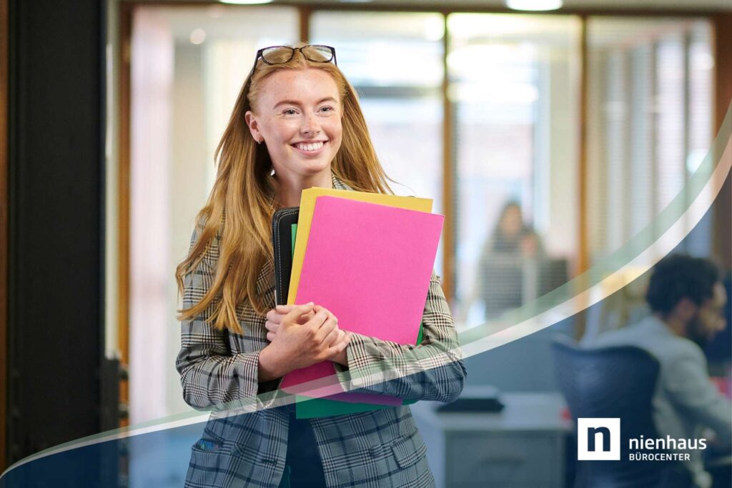 Eine junge rothaarige Frau mit Ordnern in der Hand läuft lachend und glücklich durch ein Büro.