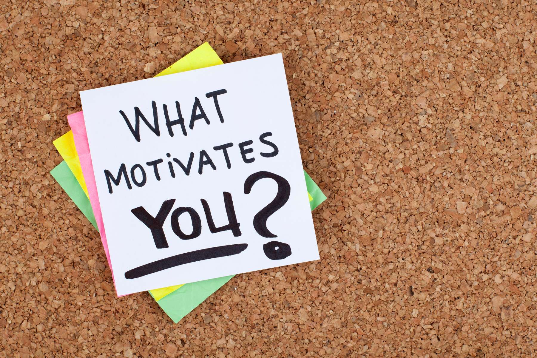 Auf einer Unterlage aus Kork liegen verschiedenfarbige Zettel, während auf dem obersten Zettel die Frage What motivates you? geschrieben steht.