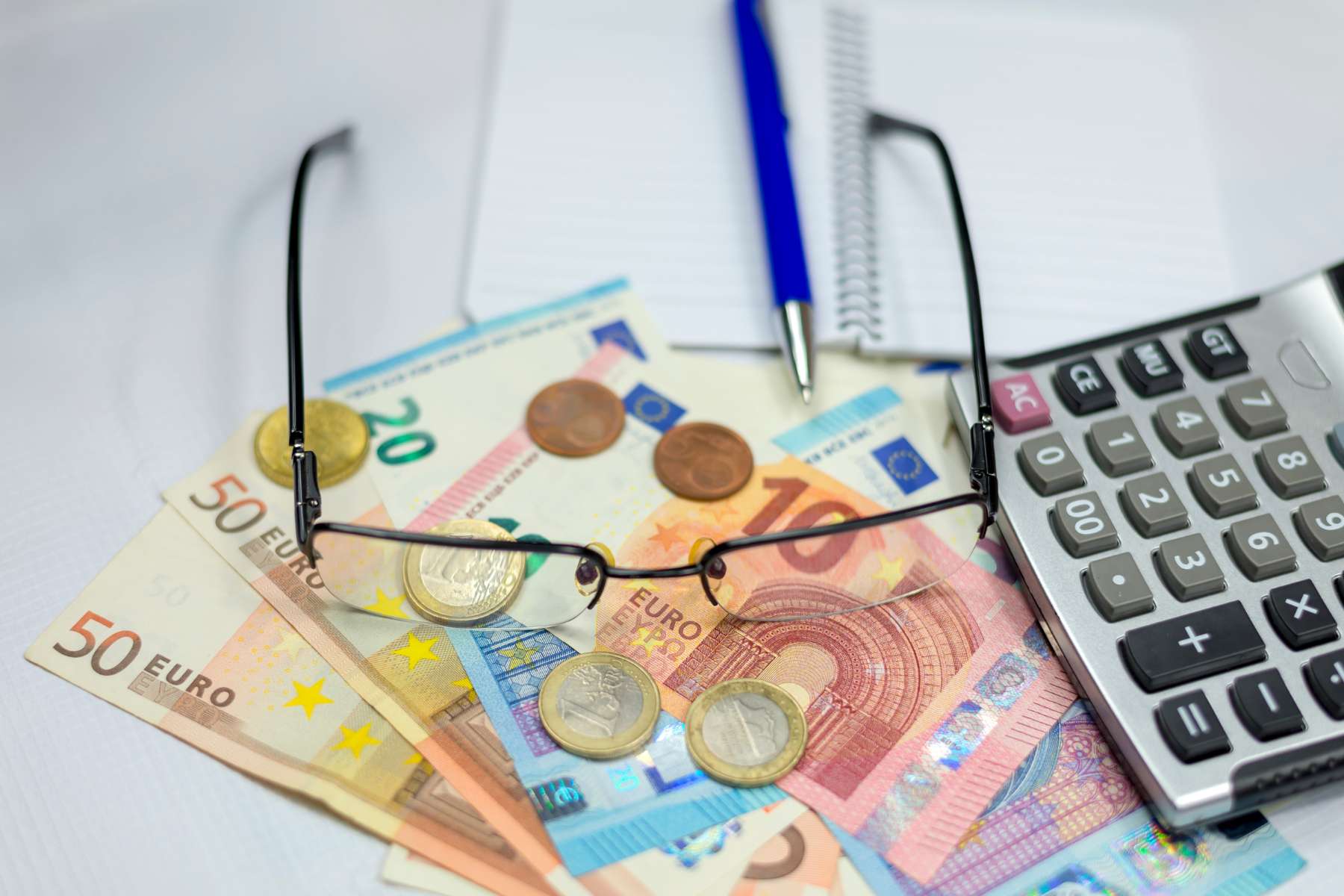 Nahaufnahme von Geldscheinen und Münzen, die zusammen mit einer Brille, einem Kugelschreiber und einem Taschenrechner auf einem Schreibtisch liegen.