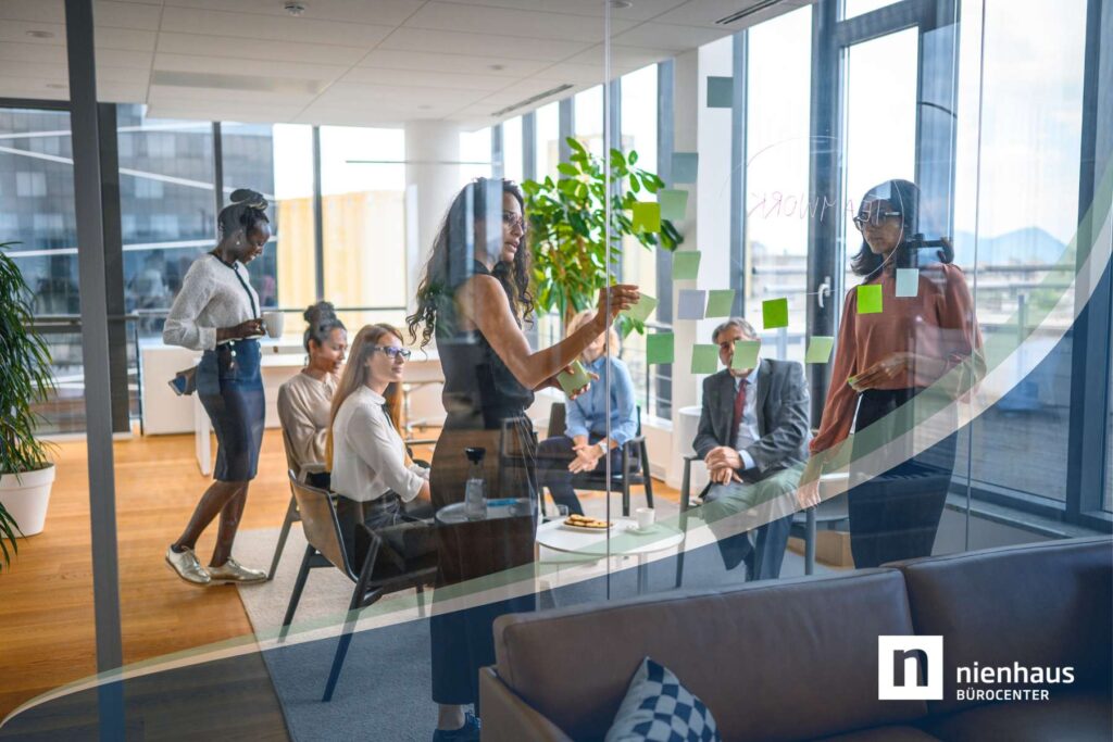 Eine Gruppe Angestellter sitzt in einem abgetrennten Büroraum im Kreis während Führungskräfte an einer Scheibe mit Post-Its stehen und etwas erklären.