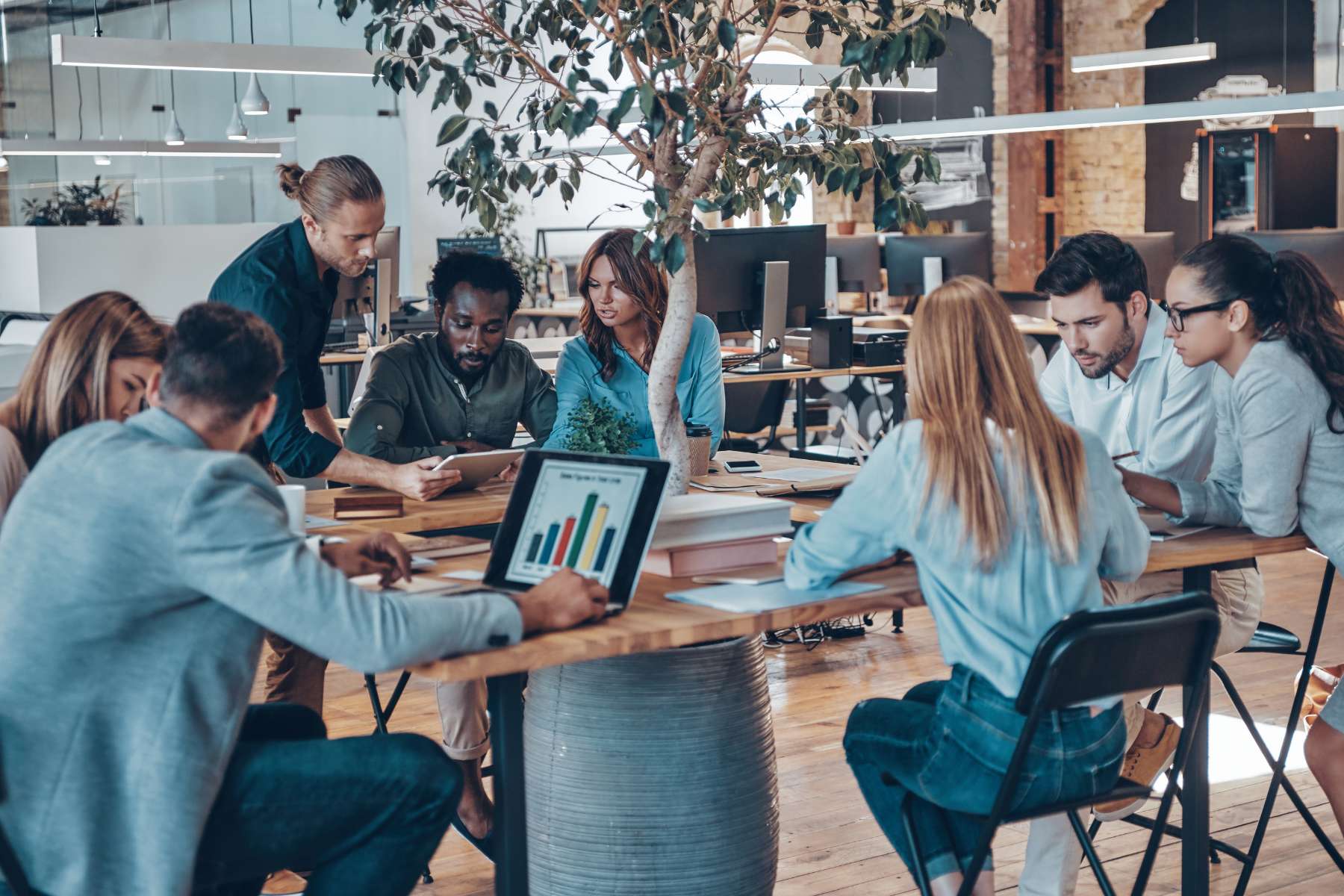 Eine Gruppe diverser Büroangestellter sitzt an einem viereckigen Holztisch, arbeitet an Laptops und unterhält sich miteinander, während eine männliche Führungskraft zwei Mitarbeitern etwas auf einem Tablet zeigt.