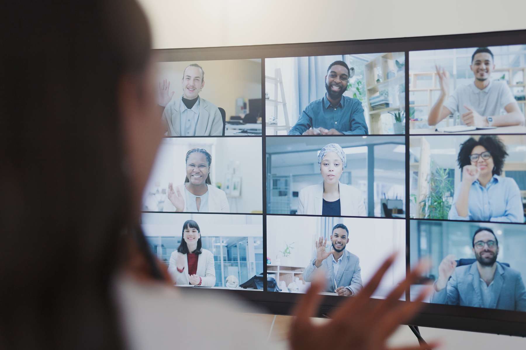 Eine weibliche Führungskraft sitzt in einem Videocall vor einem Bildschirm, auf dem 9 lächelnde Teilnehmer zu sehen sind und begrüßt diese mit einem Winken.