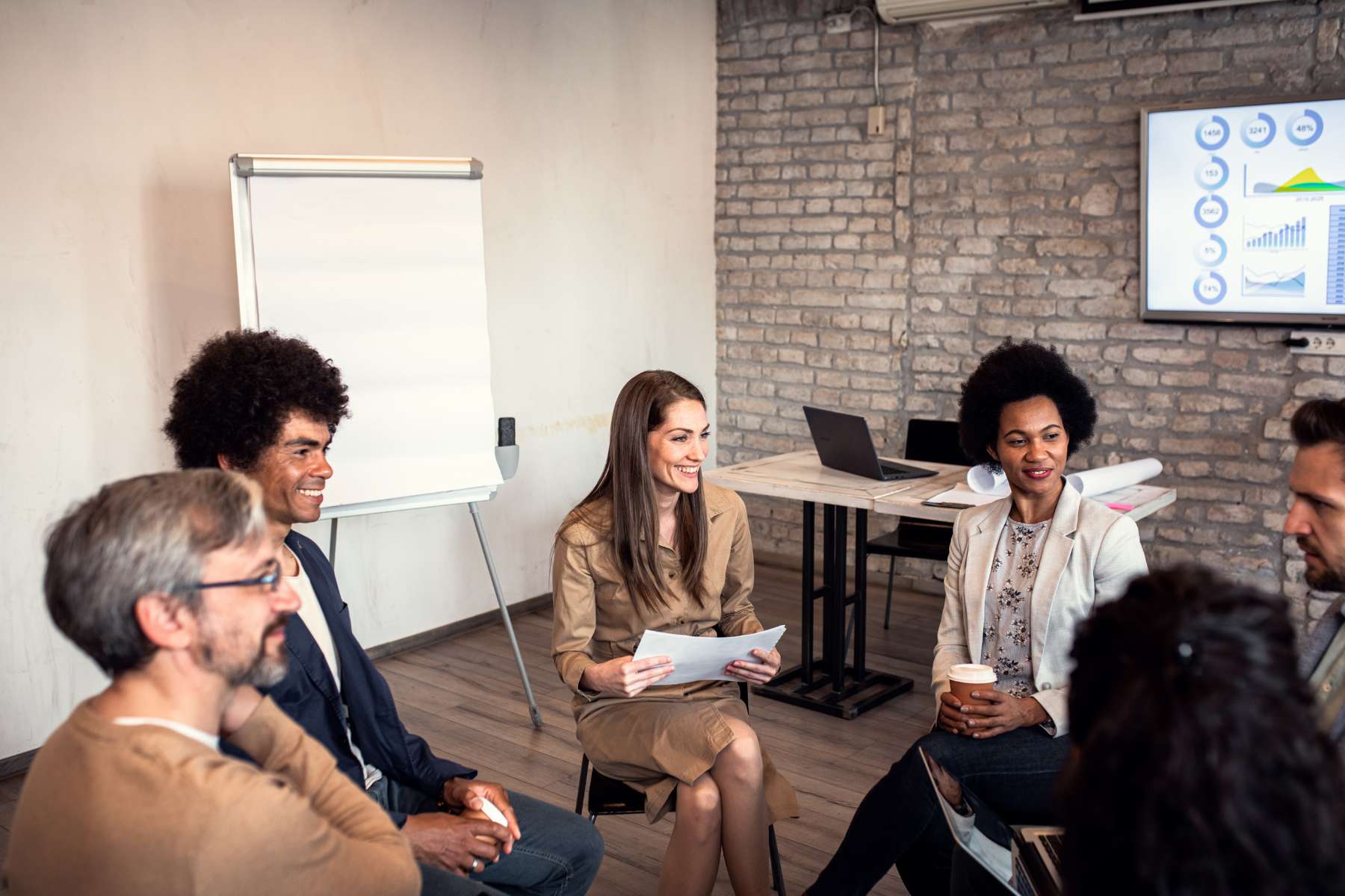 Eine Gruppe diverser Angestellter sitzt im Kreis in einem Büro und hört lächelnd einer Führungskraft zu.