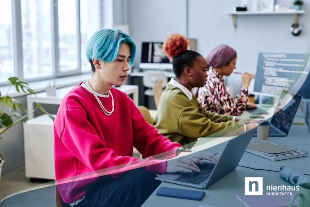 Ein junger Mann und zwei junge Frauen der Gen Z sitzen im Büro an Schreibtischen und arbeiten konzentriert an Laptops und Monitoren.