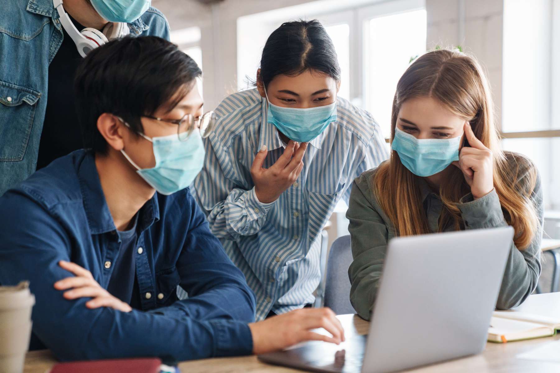 Junge Menschen der Generation Z arbeiten während der Corona-Pandemie im Büro am Laptop, tragen Schutzmasken und unterhalten sich.