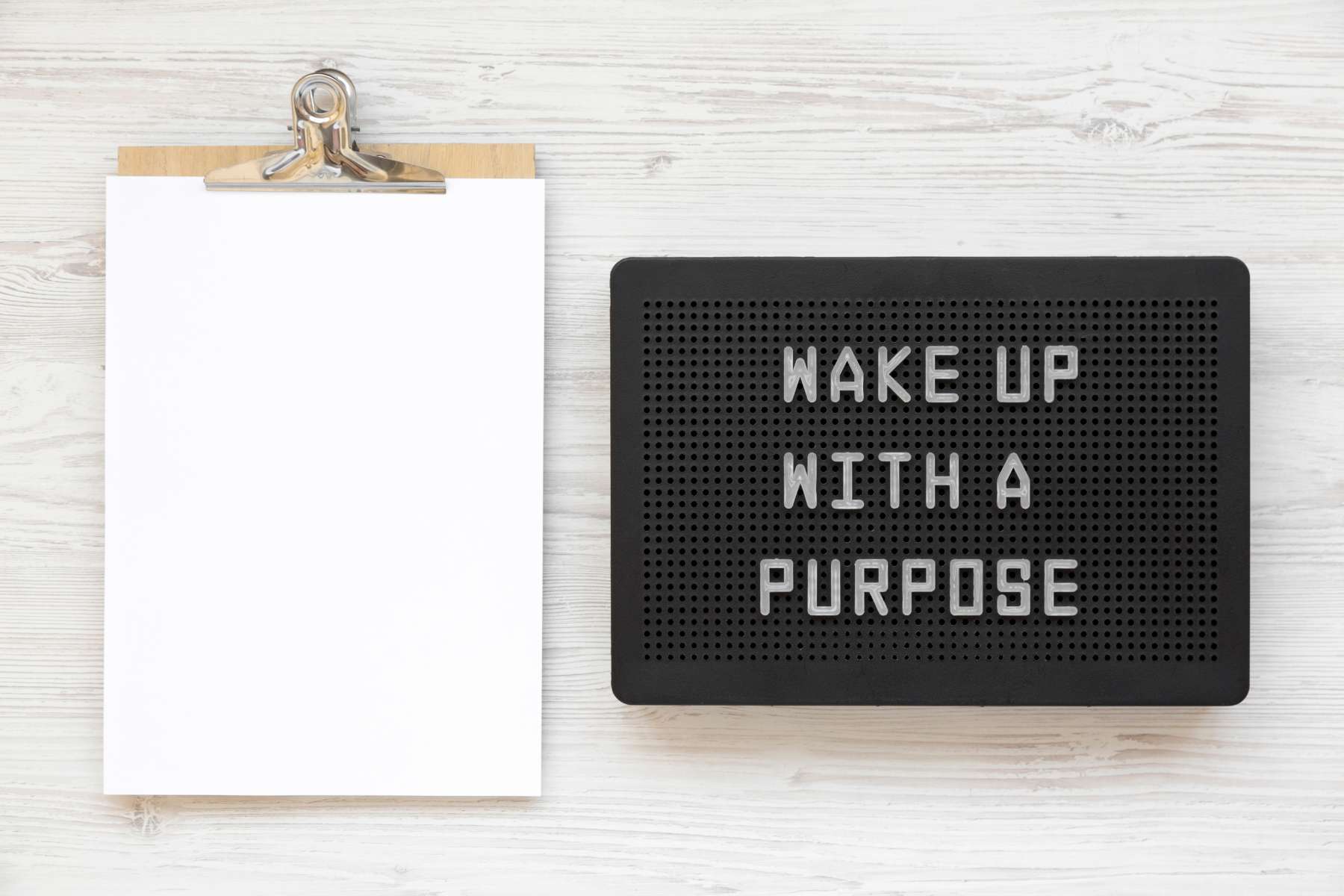 Auf einem hellen Holztisch liegt ein Klemmbrett mit weißen Zetteln und ein schwarzes Display auf dem der Satz wake up with a purpose abgebildet ist.