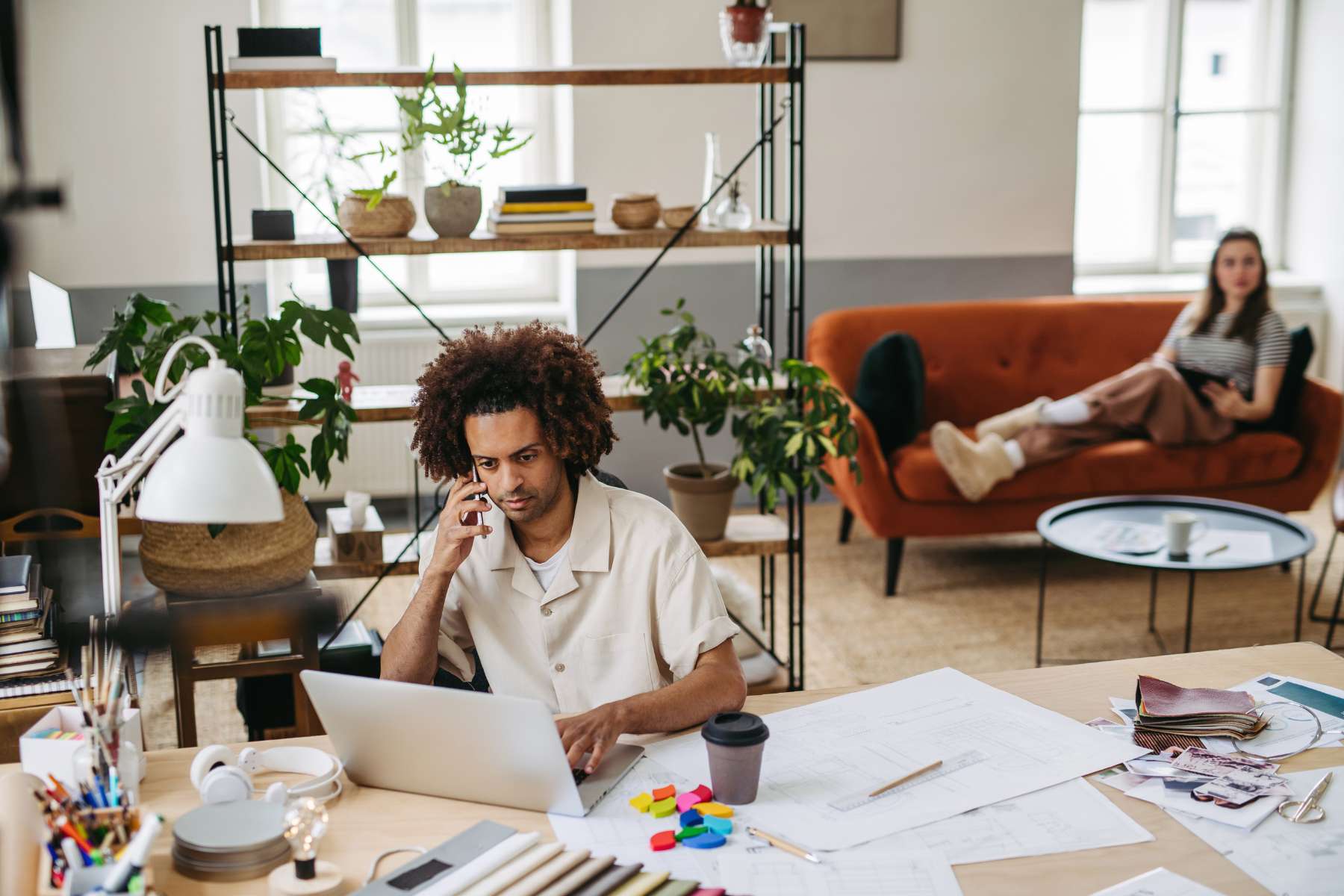 Ein junger Mitarbeiter der Generation Z sitzt telefonierend an einem Schreitisch und arbeitet am Laptop, während im hinteren Teil des Büros eine junge Kollegin auf einem Sofa sitzt und am Tablet arbeitet.