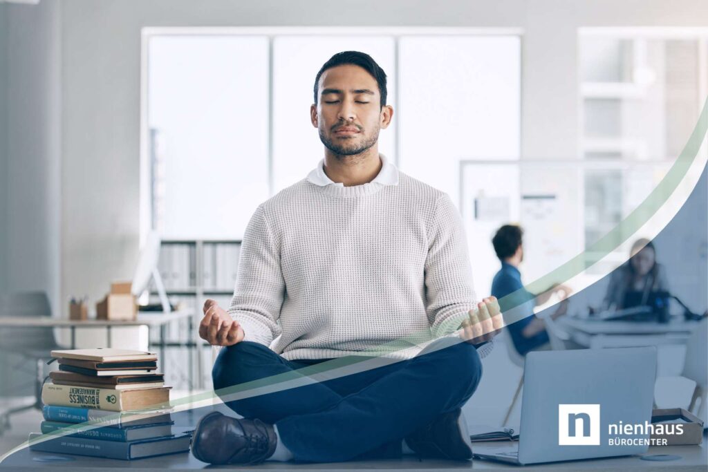 Ein männlicher Büroangestellter sitzt in Lotushaltung und mit geschlossenen Augen auf seinem Schreibtisch, während man im Hintergrund ein Büro und zwei Arbeitskollegen sieht, als Sinnbild für Calm Technology im Büro.