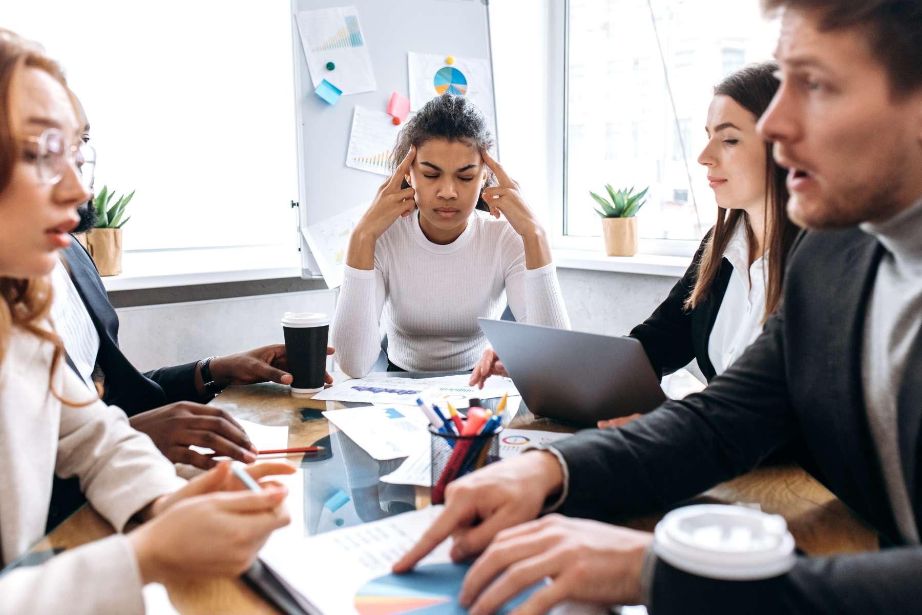 Eine junge Angestellte sitzt mit 4 Kollegen am Schreibtisch in einem Meeting und hält sich gestresst die Mittelfinger an die Schläfen, während die anderen über etwas diskutieren.