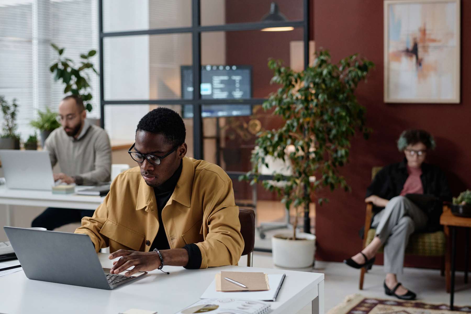 Zwei männliche und eine weibliche Büroangestellte sitzen an Schreibtischen und arbeiten konzentriert an Laptops, während im Hintergrund ein Konferenzraum mit Monitor verschwommen sichtbar ist.