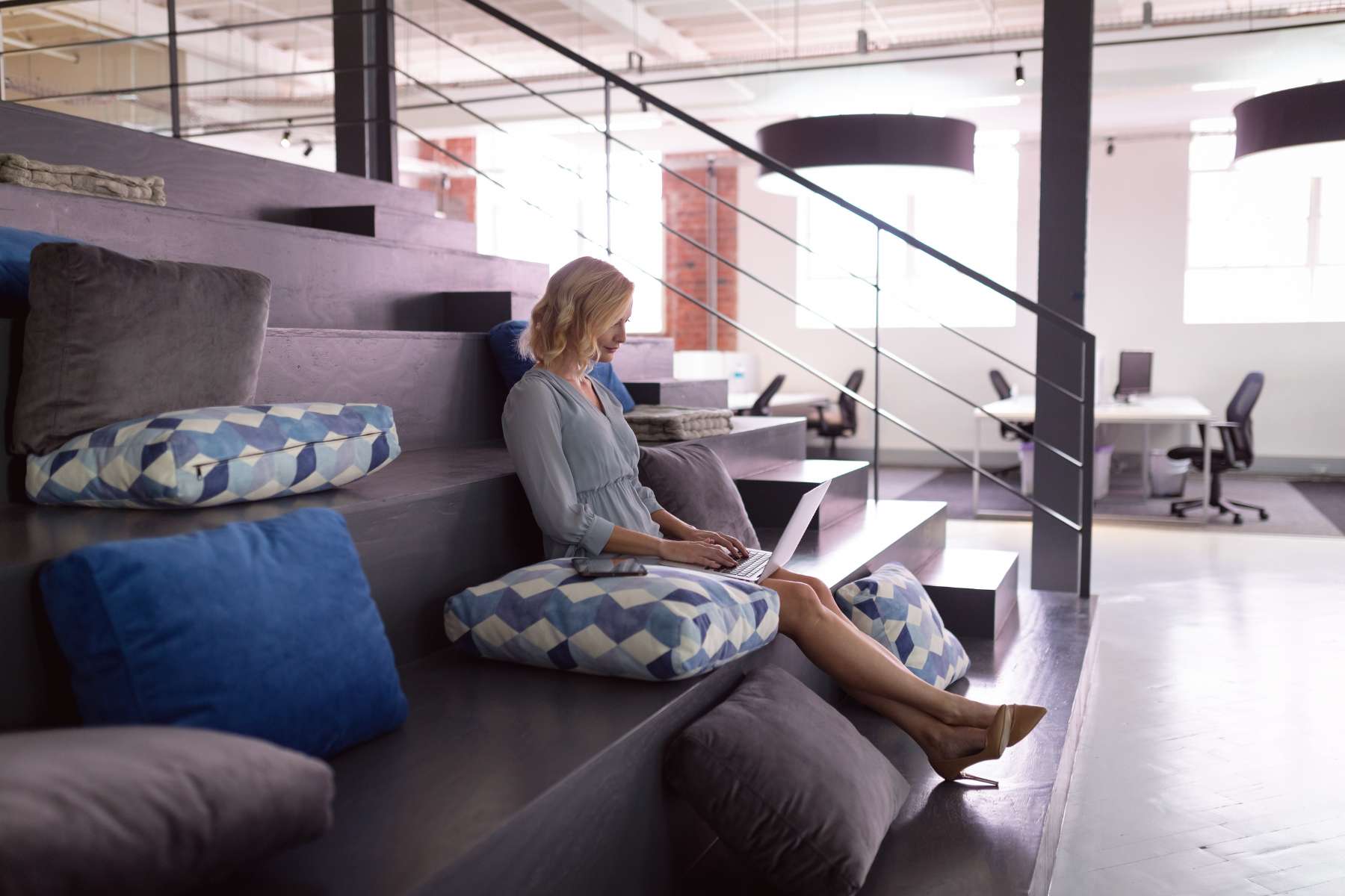 Eine weibliche Angestellte sitzt in einem großen offenen Büro auf einer Tribüne mit verschiedenfarbigen Kissen und arbeitet entspannt am Laptop.