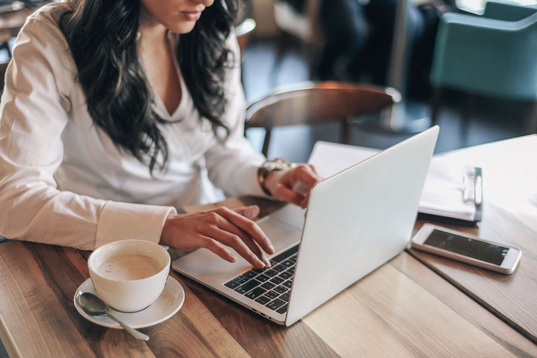 Junge Frau sitzt im Home Office to Go in einem Cafe an einem Holztisch, trinkt einen Kaffee und arbeitet am Laptop.