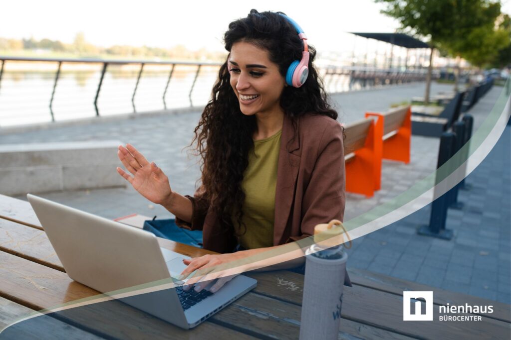 Eine junge Frau sitzt draußen an einer Promenade an einem Holztisch im Home-Office to Go lachend vor einem Laptop und hat Kopfhörer auf.