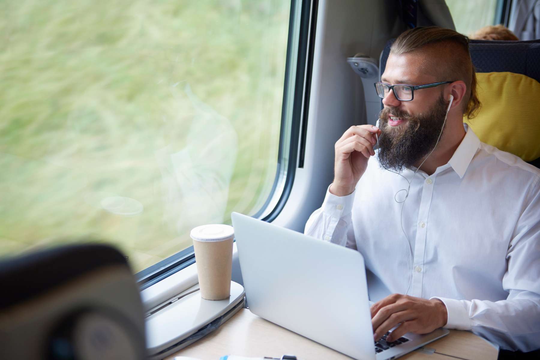 Junger Mann sitzt im Zug, arbeitet im Home -Office to Go am Laptop und telefoniert dabei mit In-Ear-Kopfhörern.