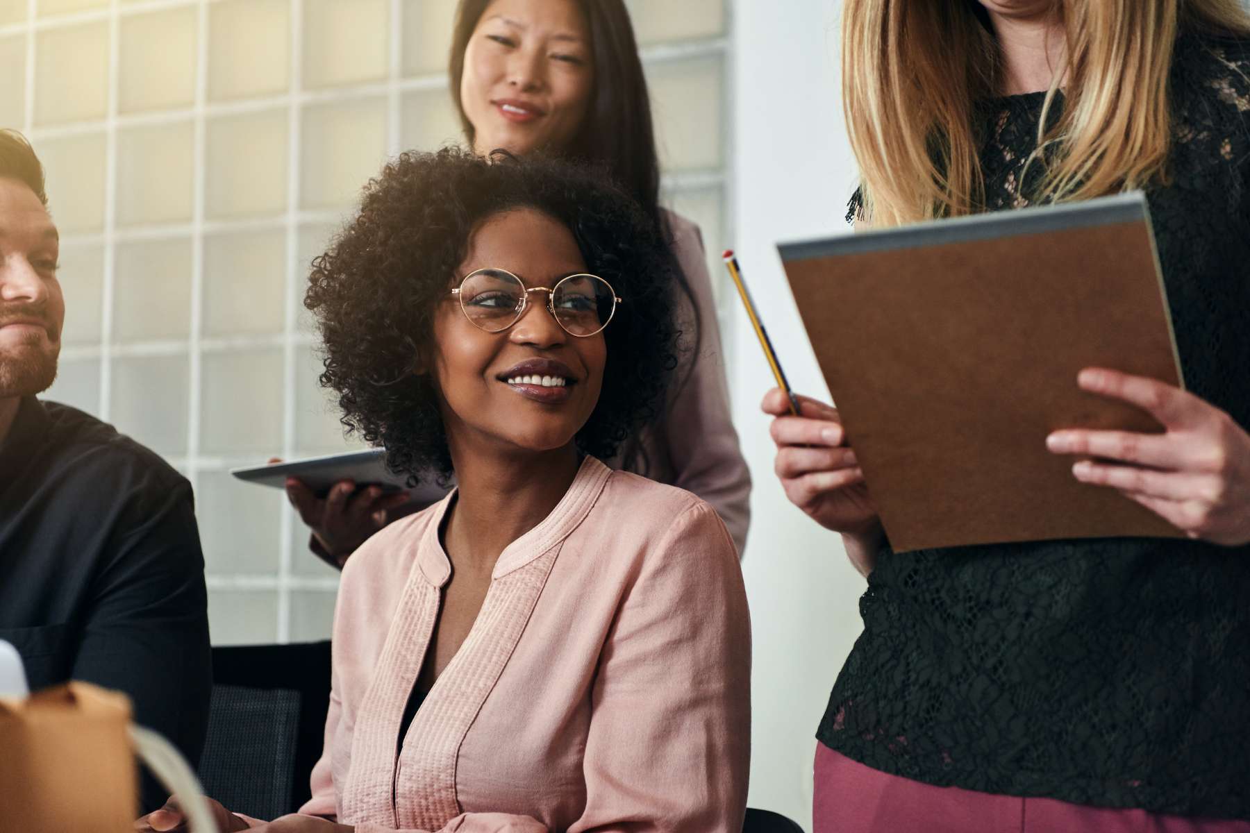 Eine schwarze Frau mittleren Alters sitzt an einem Schreibtisch, lächelt und ist von Arbeitskollegen umgeben.