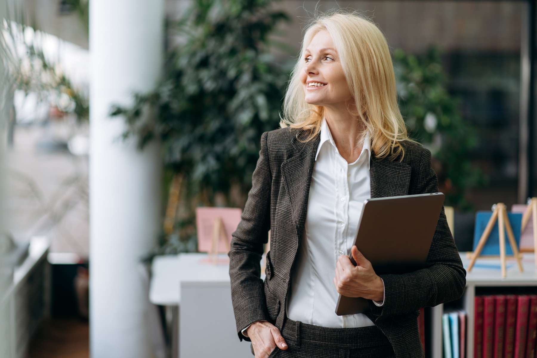 Eine mittelalte blonde Geschäftsfrau steht lächeln in einem Büro und schaut zur Seite, während sie ein Tablet im Arm hält.