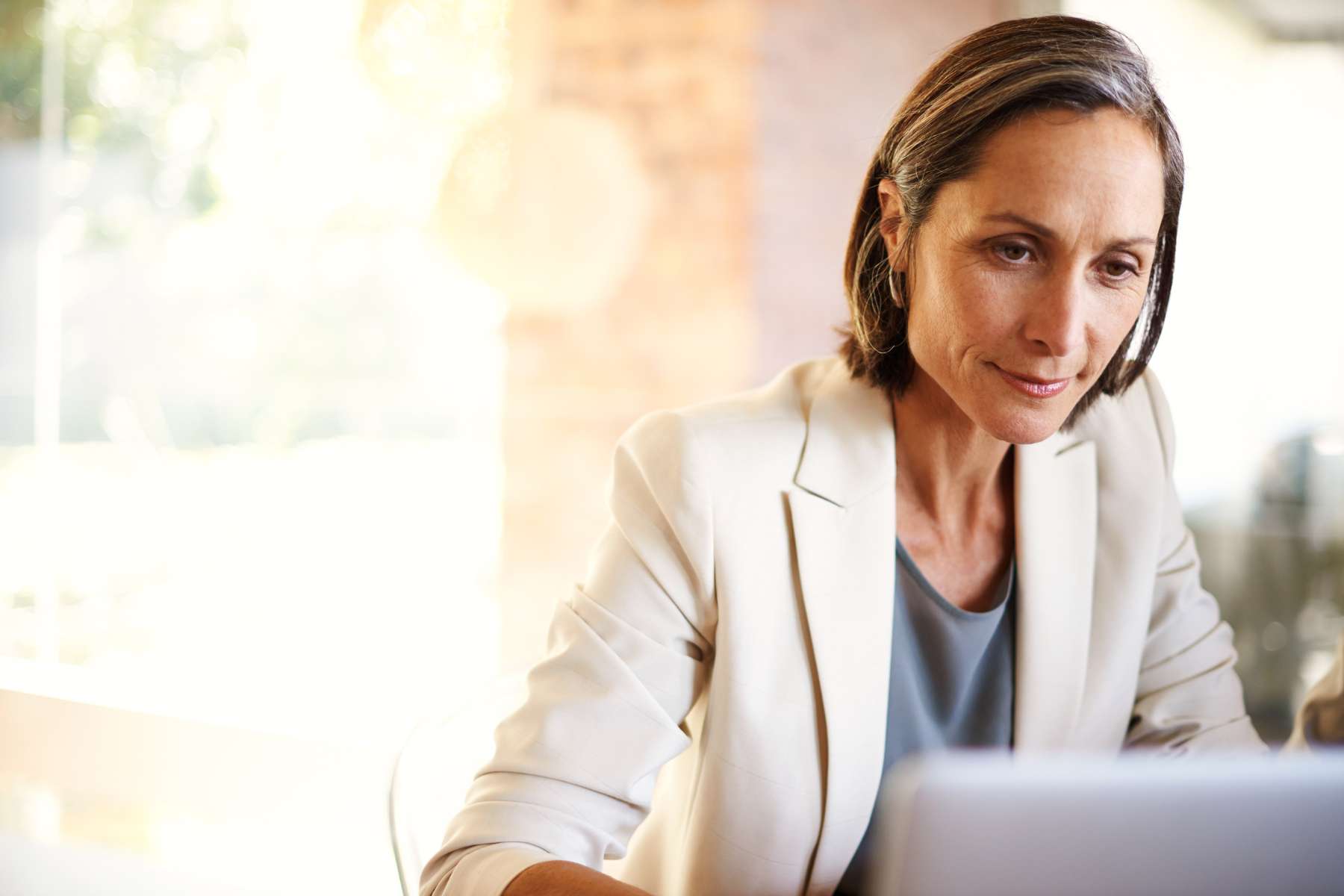 Eine mittelalte Büroangestellte im hellen Anzug sitzt an einem Schreibtisch, wirkt zufrieden und arbeitet an einem Laptop.