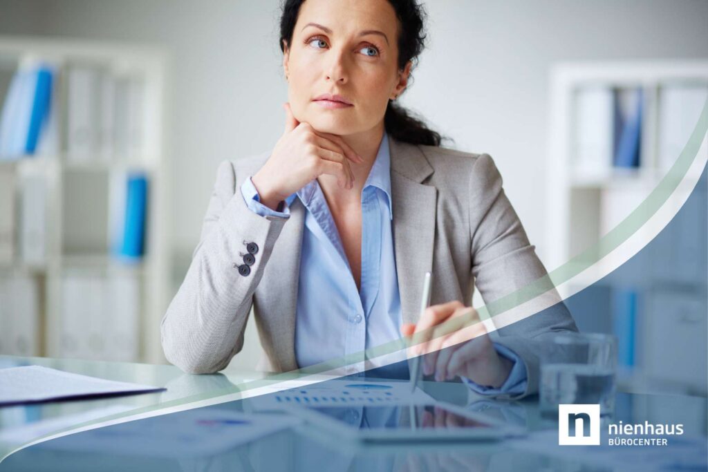 Eine mittelalte Geschäftsfrau sitzt nachdenklich an einem Schreibtisch, als Sinnbild zum Thema Menopause im Job.