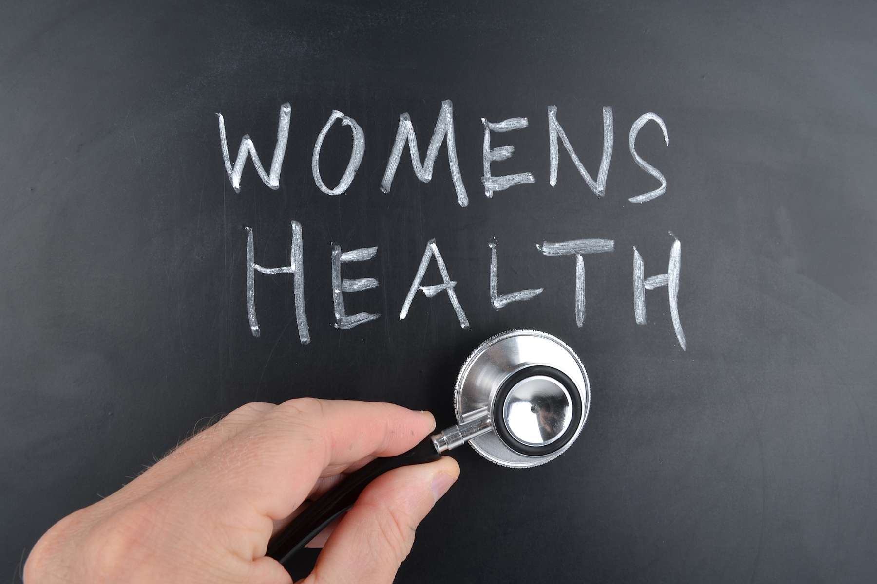 Ein Stethoskop wird von einer Hand auf eine schwarze Tafel gedrückt, auf der in Weiß die Worte Womens Health stehen.