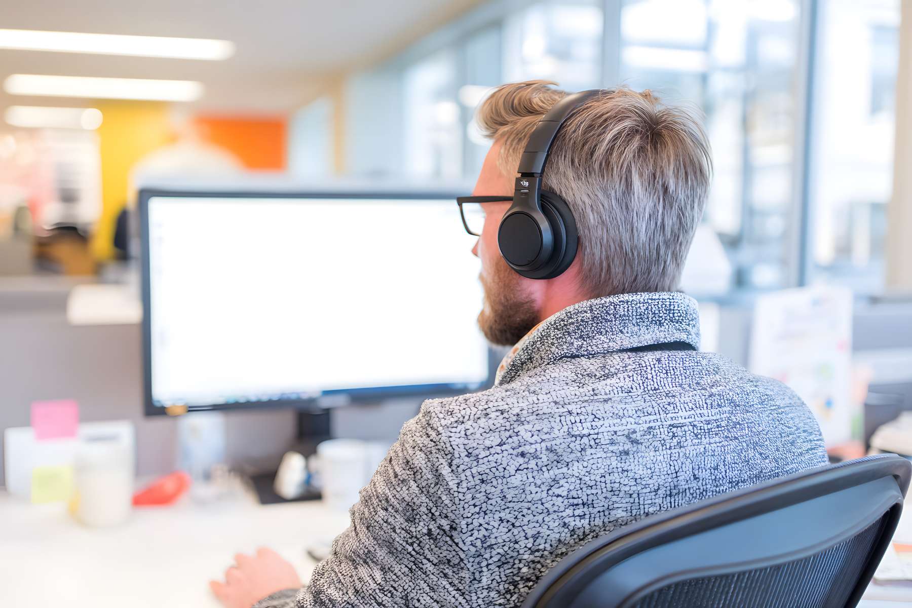 Ein Mann in grauem Pullover und mit grauen Haaren sitzt mit Kopfhörern an einem Schreibtisch im Büro und arbeitet an einem PC.